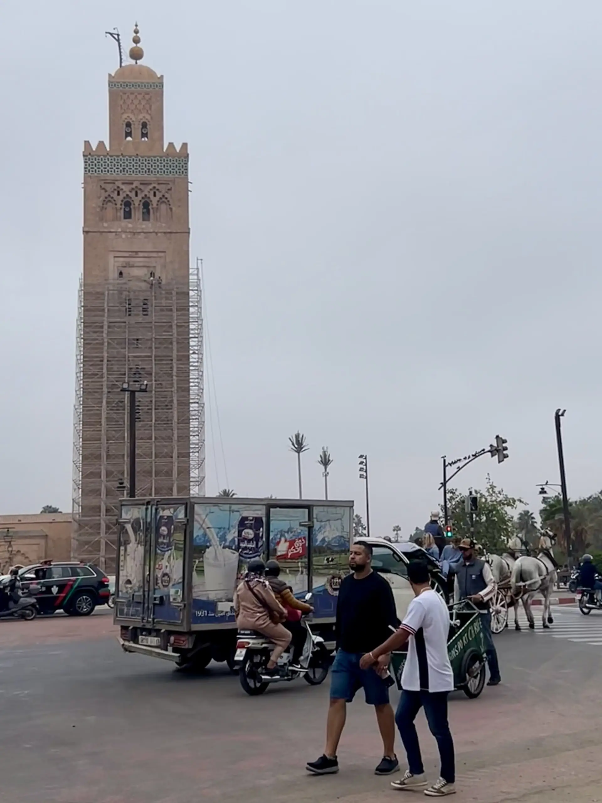 Minaret mešity Koutoubia obalený lešením se tyčí nad rušnou křižovatkou s nákladním autem, motorkami a koňským kočárem.