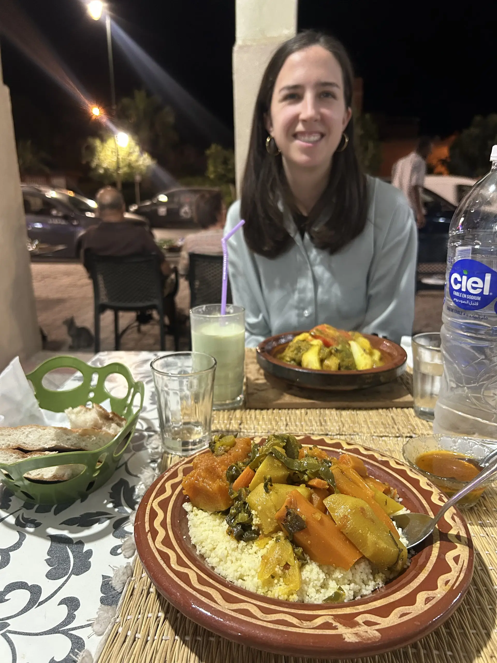 Anička sedí na večerní terase restaurace, na stole tažin se zeleninou na kuskusu, chléb a láhev vody.