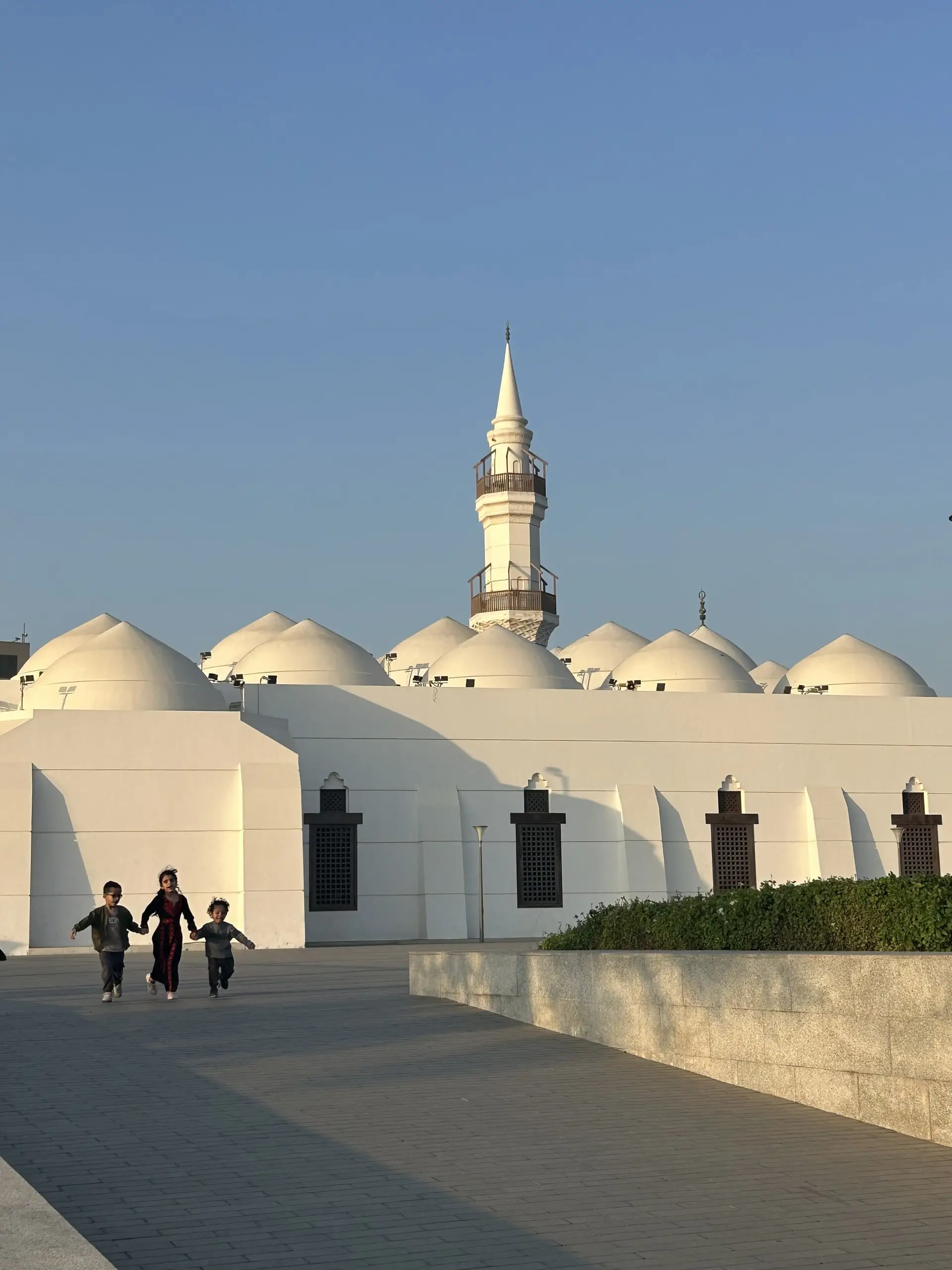 Bílá mešita s mnoha kupolemi a minaretem, před ní po prostranství běží děti.