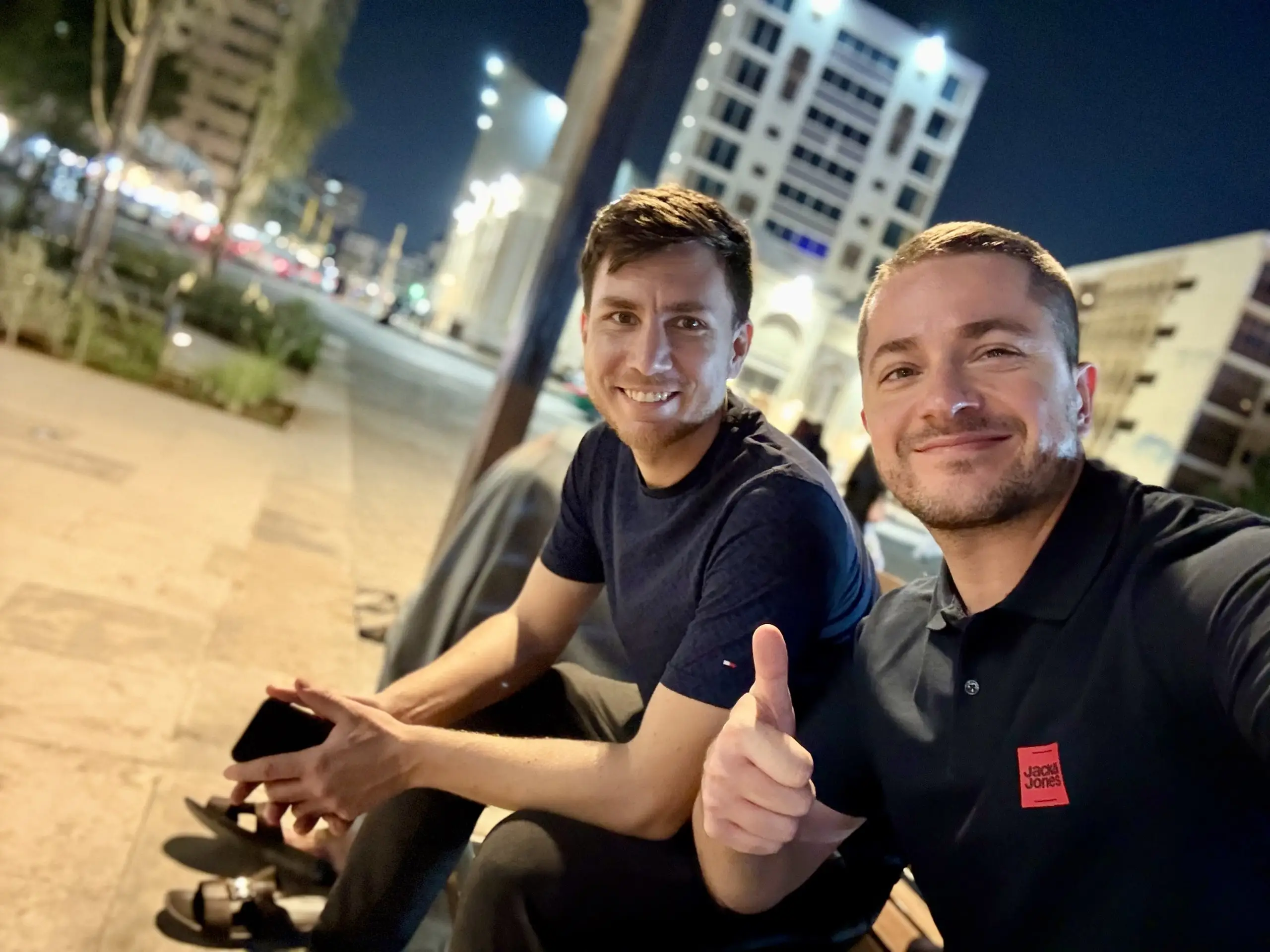 Stojím vpravo s palcem nahoru vedle kamaráda na lavičce, za námi noční osvětlené budovy.