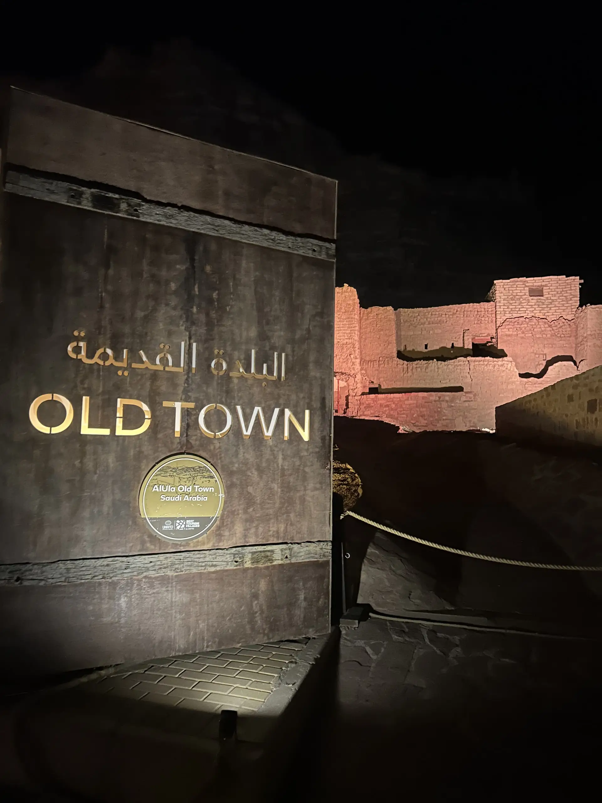 Nasvícená tabule s nápisem Old Town a arabsky, vzadu osvětlené hliněné ruiny v noci.