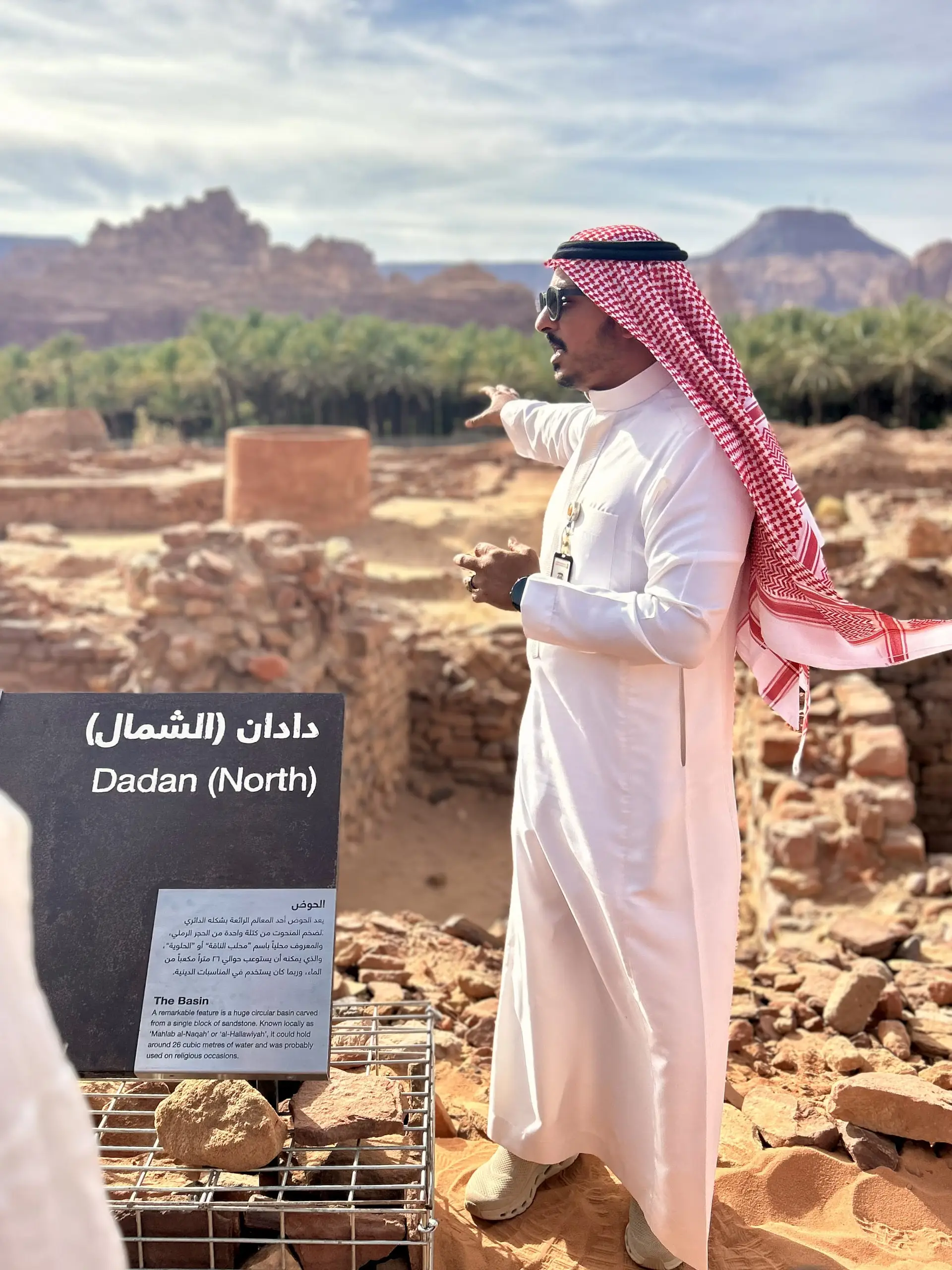 Muž v bílém thawbu a šátku vykládá u cedule Dadan (North) nad archeologickými ruinami a palmami.