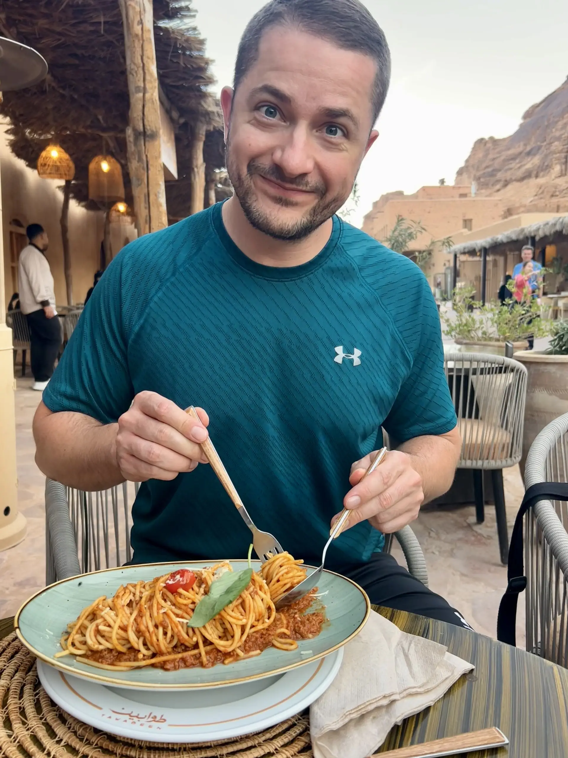 Jeden z nás se usmívá s talířem špaget na terase restaurace mezi hliněnými budovami a skalami.