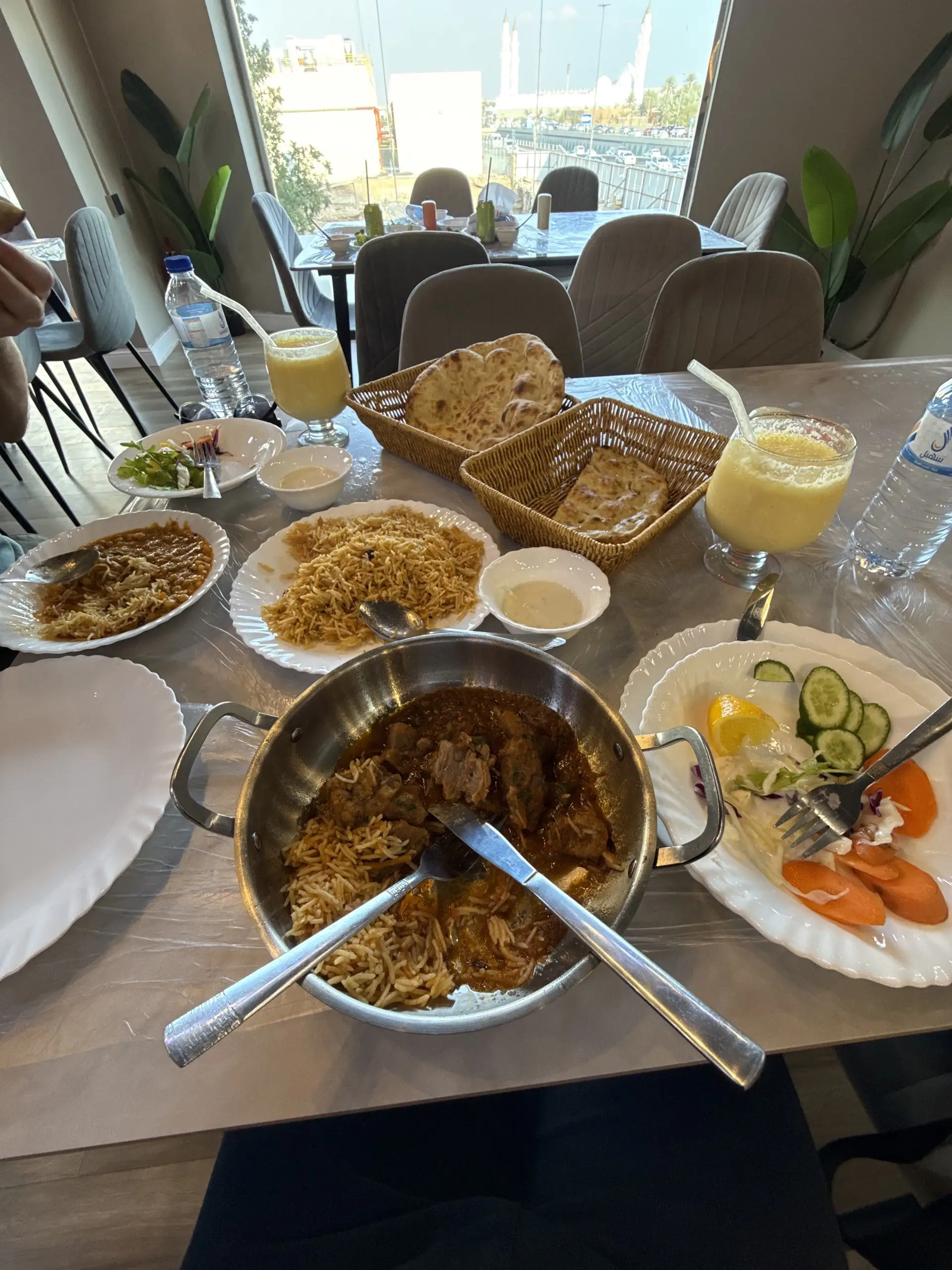 Stůl plný indicko-pákistánských jídel: kari s masem v pánvi, biryani, naan v košících, salát a dvě sklenice mango lassi, za oknem minarety.