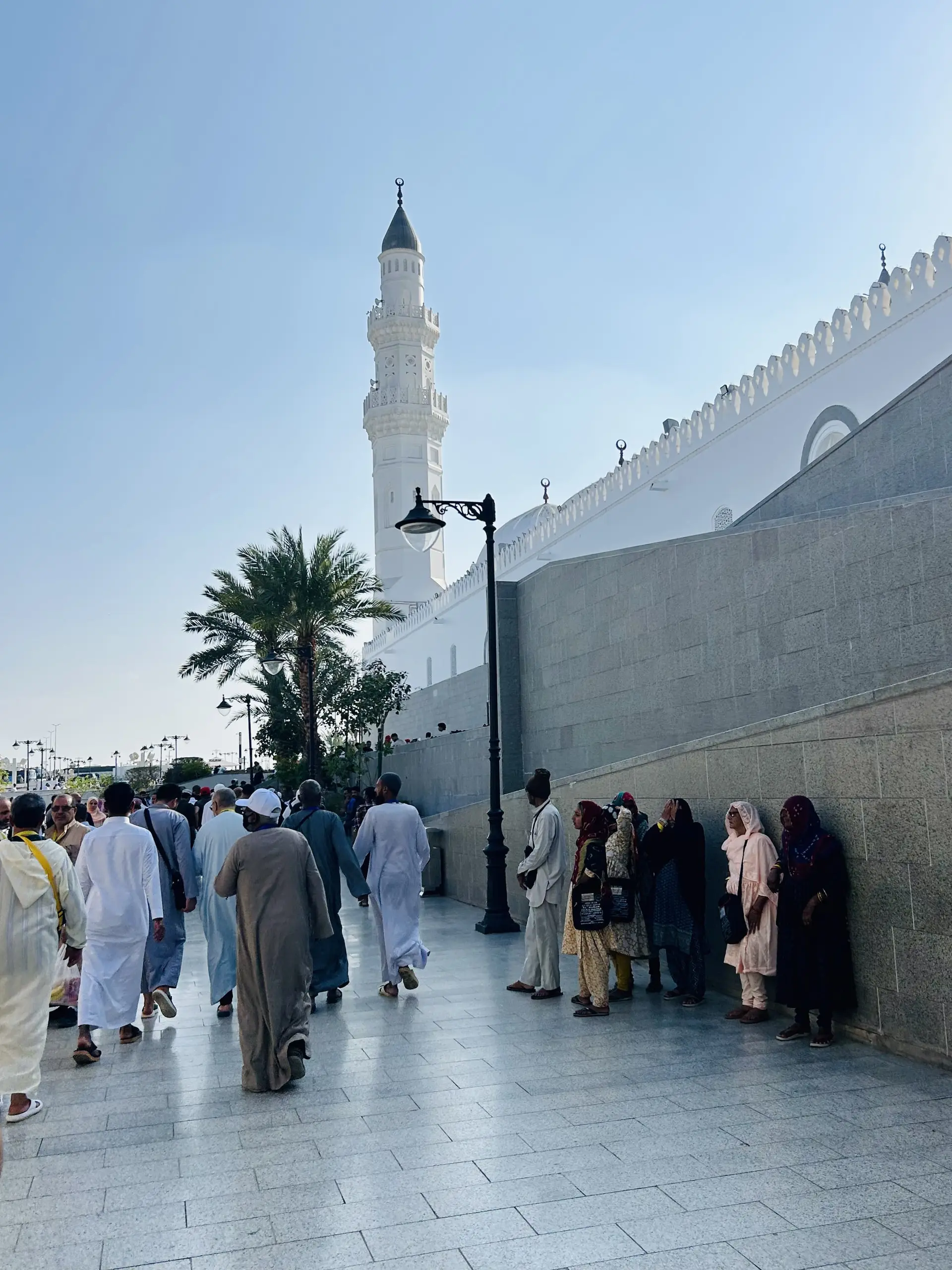 Bílá mešita Quba se štíhlým minaretem, podél zdi proudí davy poutníků v bílém oděvu, vlevo palma a pouliční lampa.