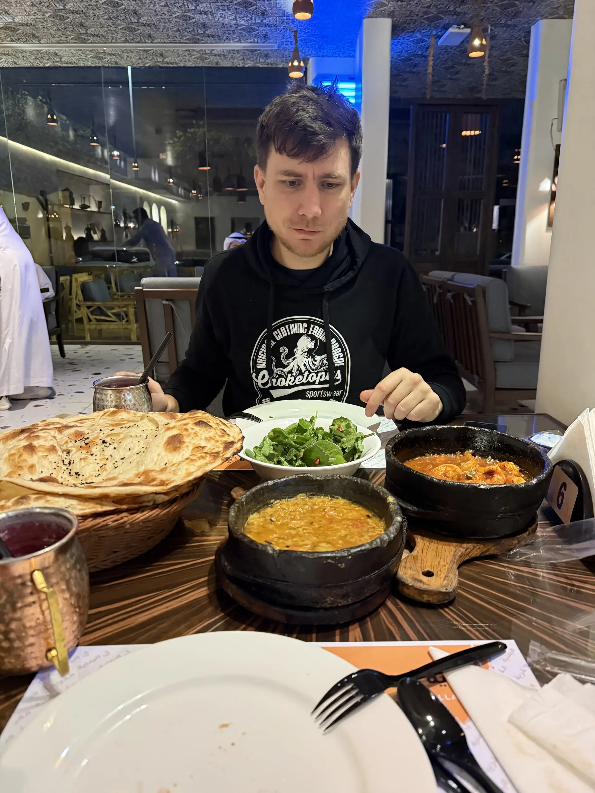 Martin u stolu jí kari z černých kamenných mističek s naanem a salátem, kolem měděné hrnečky a příbory, v pozadí osvětlený interiér restaurace.