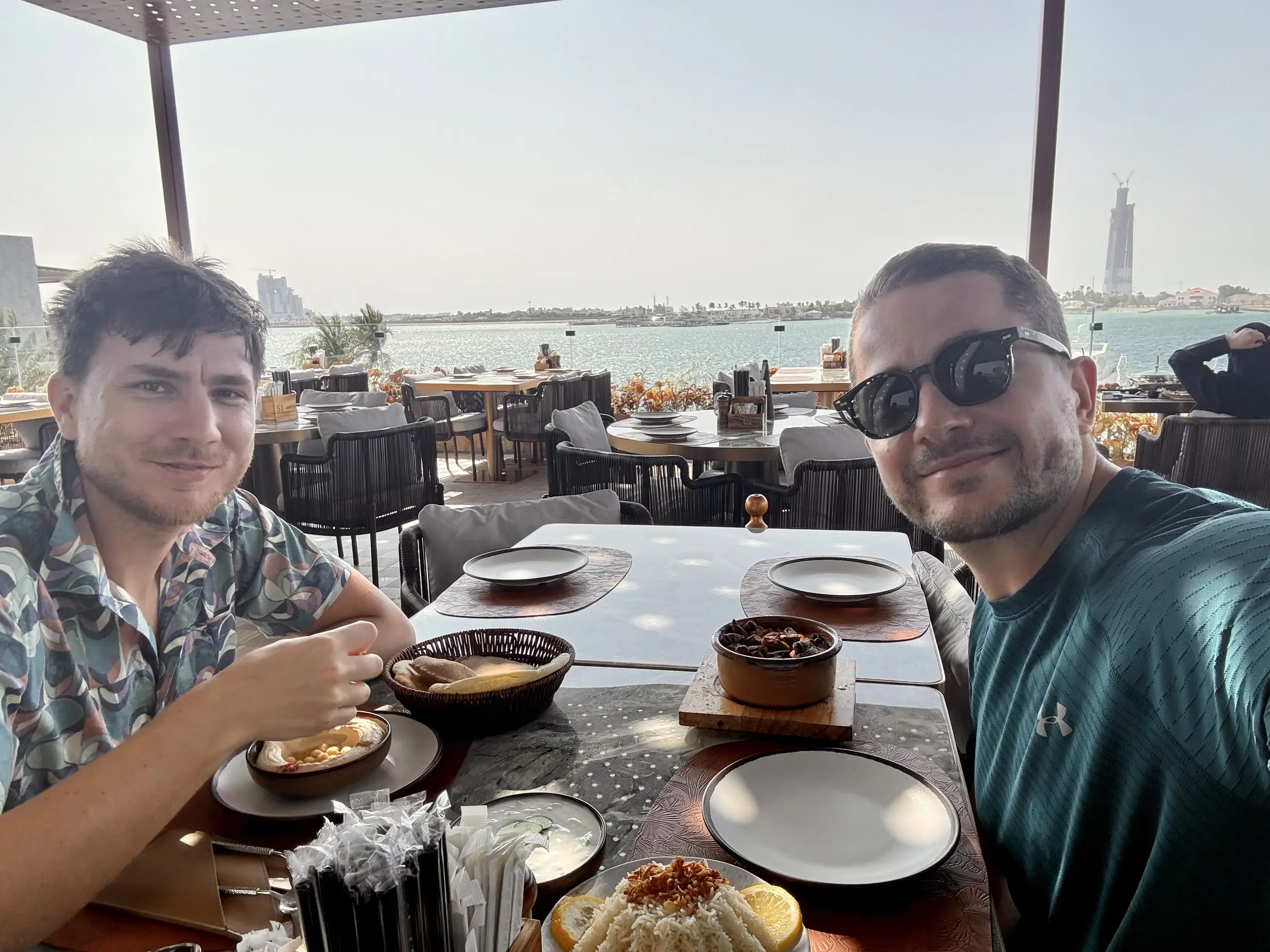 Usmíváme se do kamery u stolu s arabskými předkrmy na terase přímořské restaurace, za námi moře a mrakodrap v dálce.