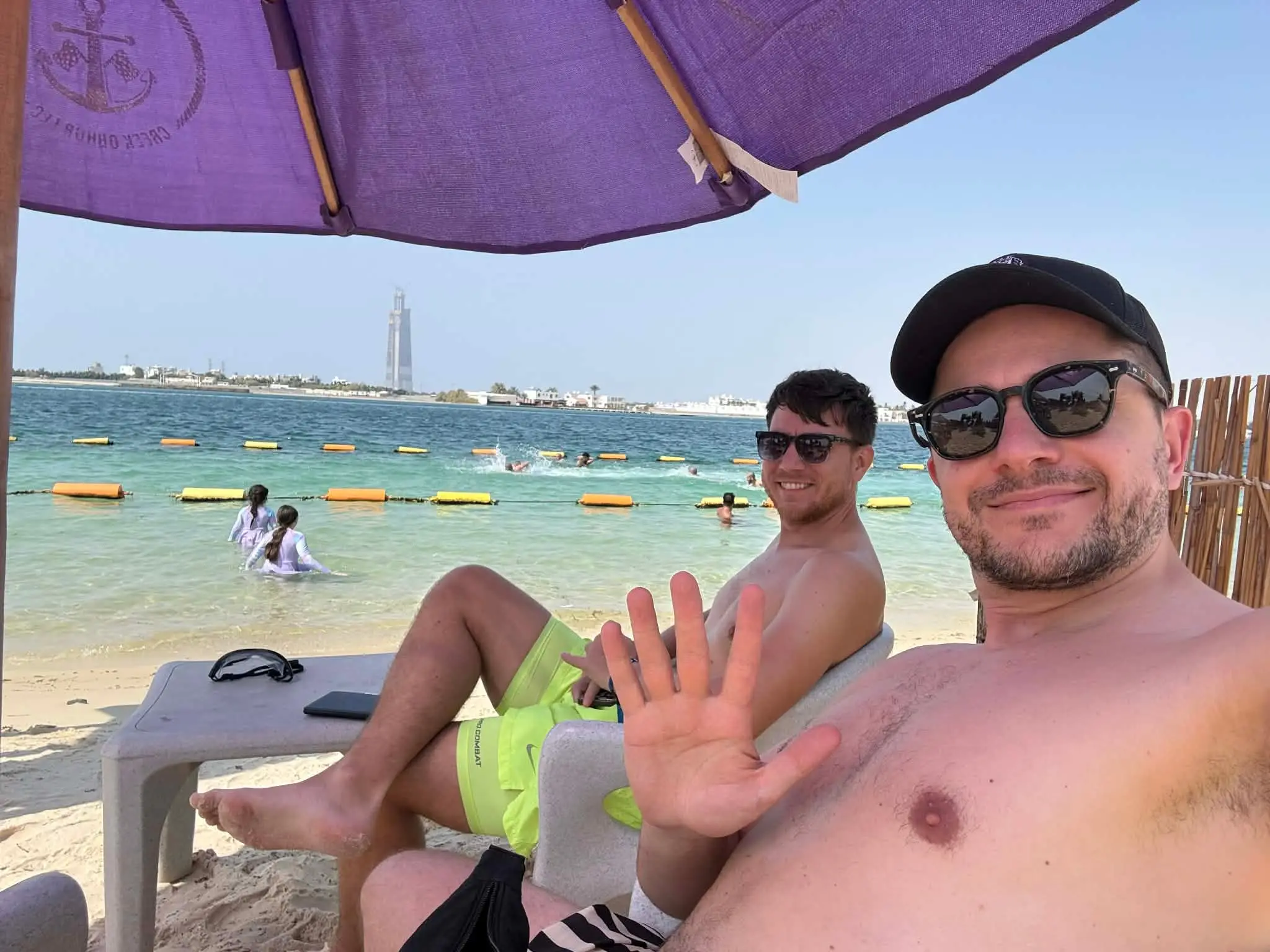 Naše selfie pod fialovým slunečníkem na pláži u tyrkysového Rudého moře, za námi se koupou lidé a na protějším břehu stojí věž.
