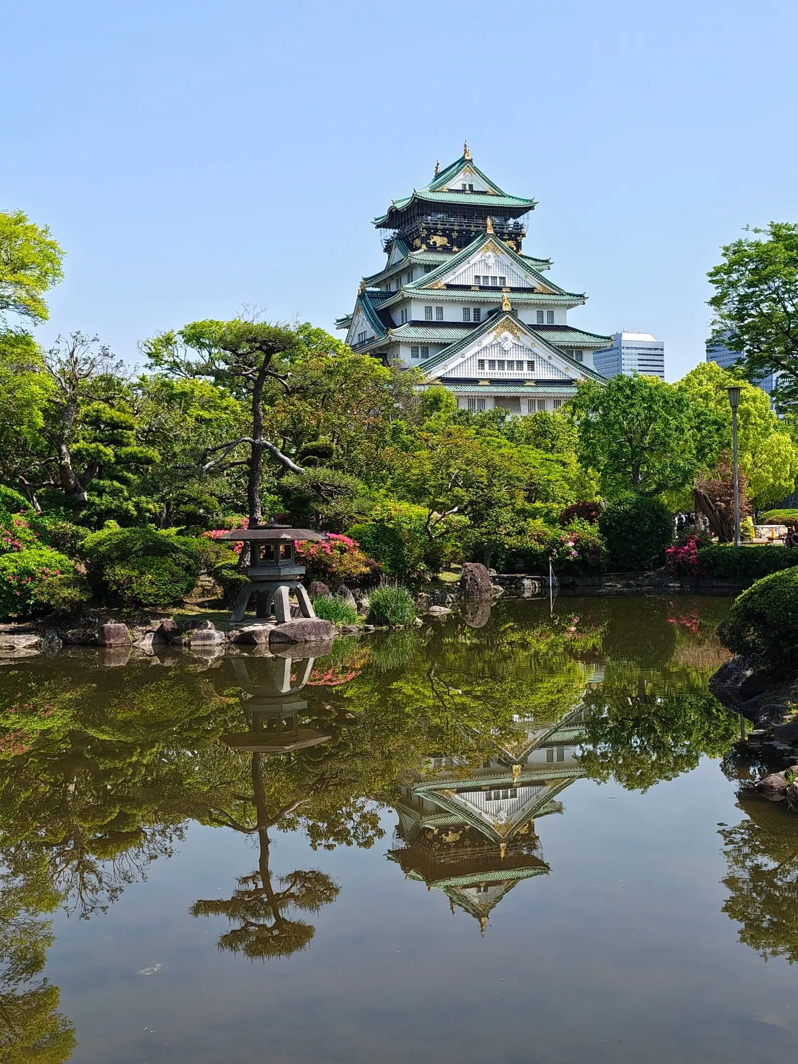 Ósacký hrad s tmavou střechou se zrcadlí v klidné hladině jezírka v japonské zahradě s tvarovanými keři.