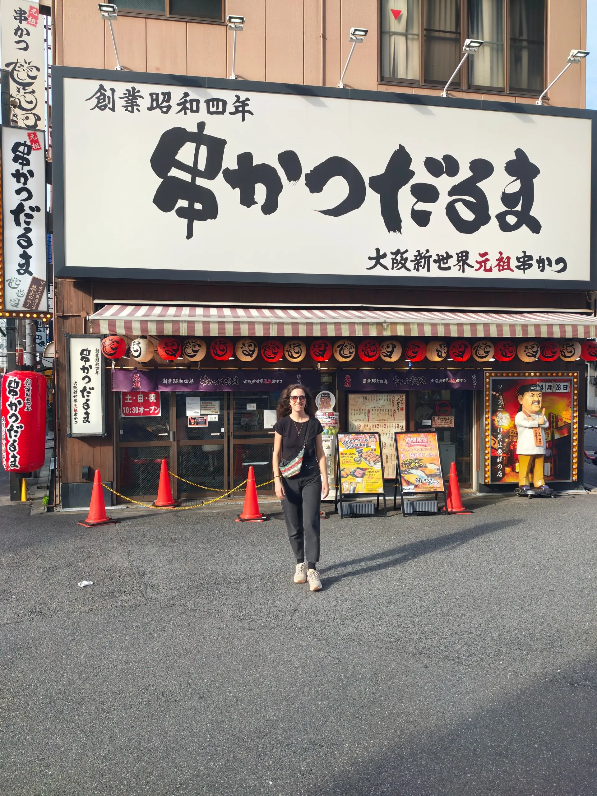 Žena jde po ulici před restaurací s velkým japonským nápisem a červenými dopravními kuželi u vchodu.