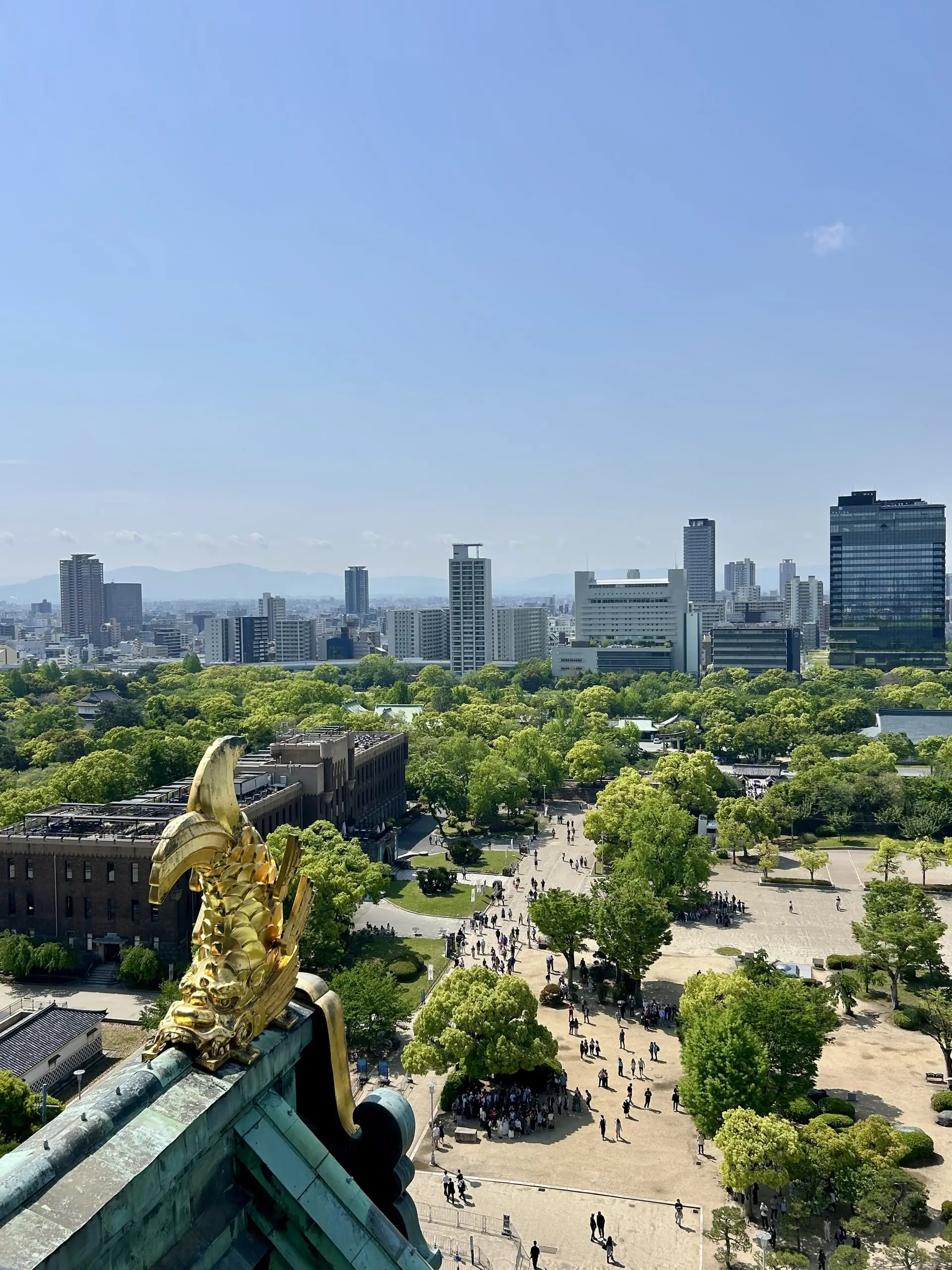 Zlatá ryba šači na okraji hradní střechy nad rozlehlým parkem a panoramatem moderních ósackých mrakodrapů.