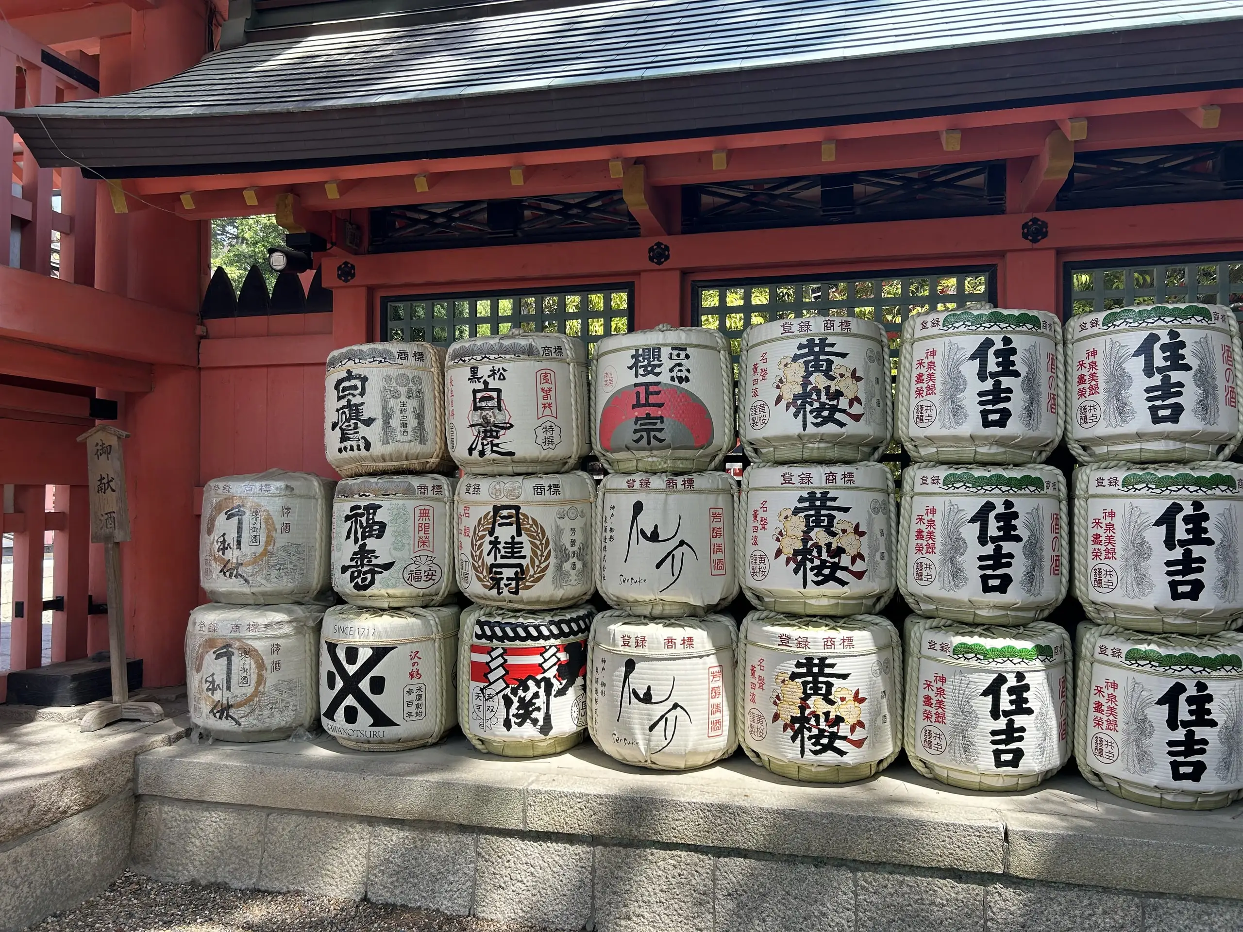 Dvě řady darovaných sudů se saké s japonskými nápisy stojí před červenou budovou šintoistické svatyně.