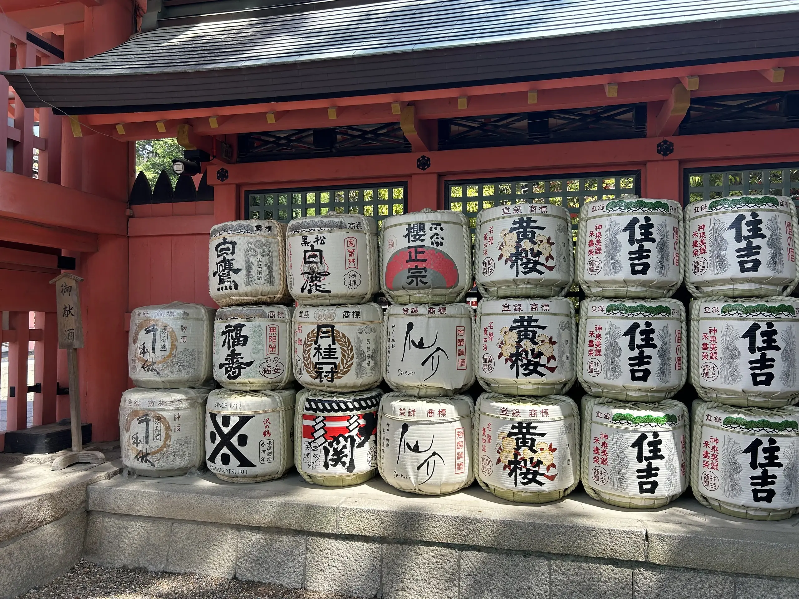 Dvě řady darovaných sudů se saké s japonskými nápisy stojí před červenou budovou šintoistické svatyně.