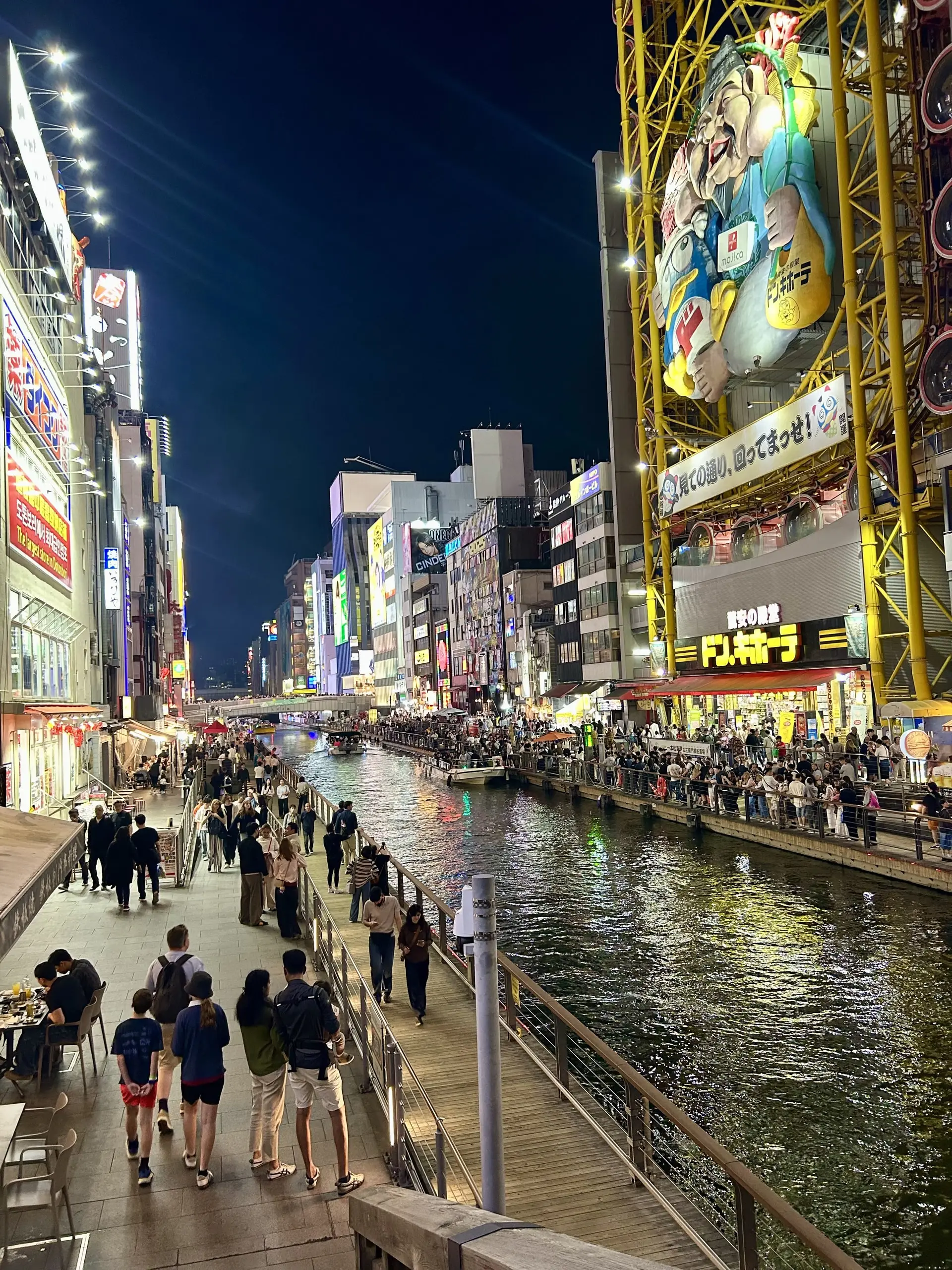Lidé se procházejí po nábřeží kanálu v Dotonbori v noci, kolem se odrážejí svítící billboardy a reklamy.