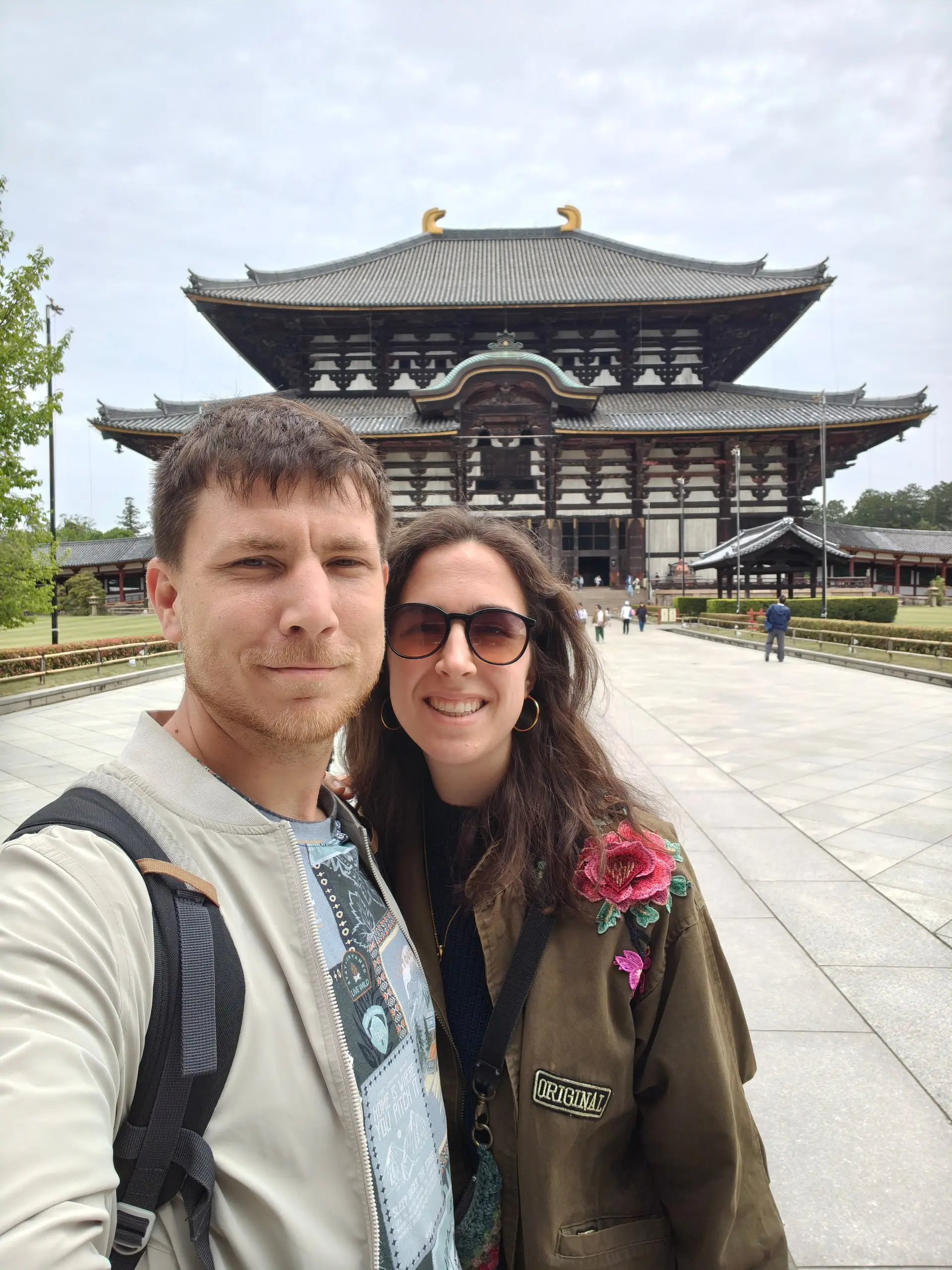 Pár se fotí na selfie před velkou dřevěnou halou chrámu Tódaidži pod zataženou oblohou.