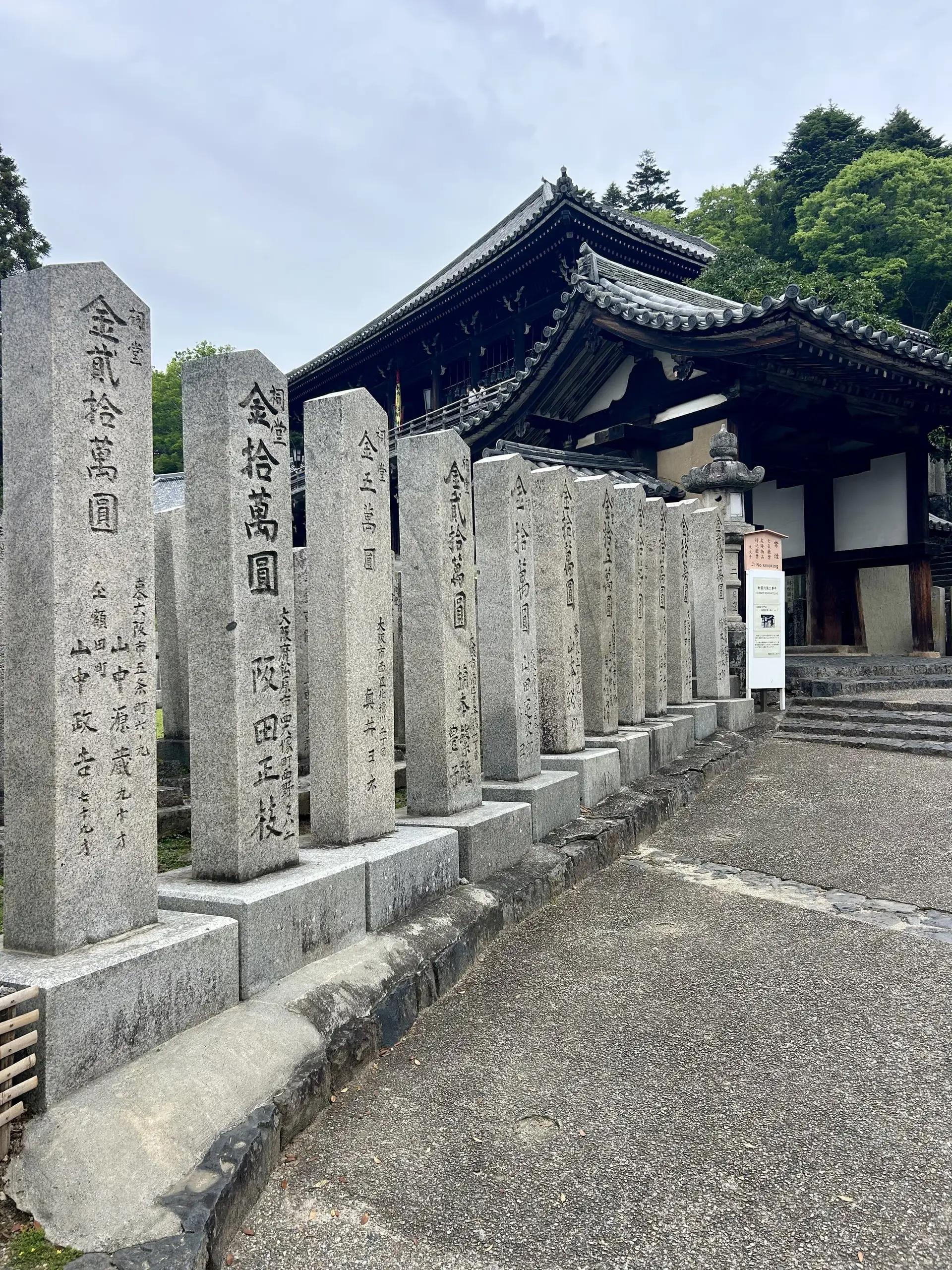 Řada vysokých kamenných stél s japonskými nápisy stojí podél cesty k dřevěné chrámové bráně.