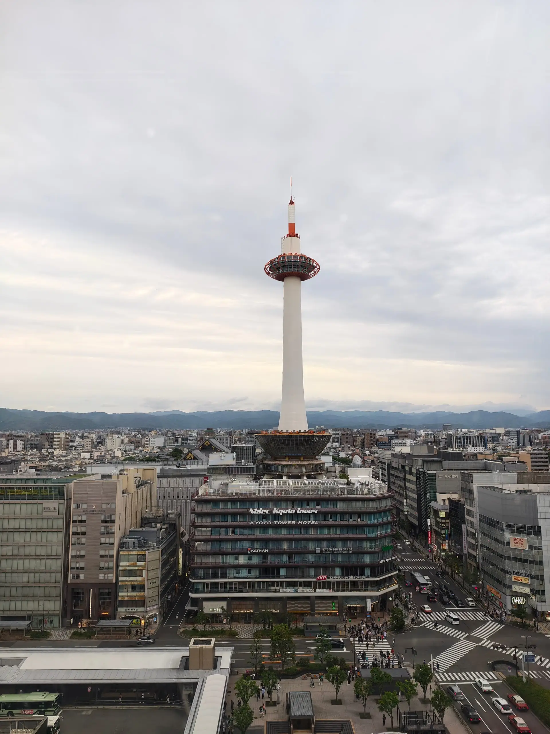 Bílá rozhledna Kjótó Tower se tyčí nad nízkou městskou zástavbou pod zataženou oblohou.