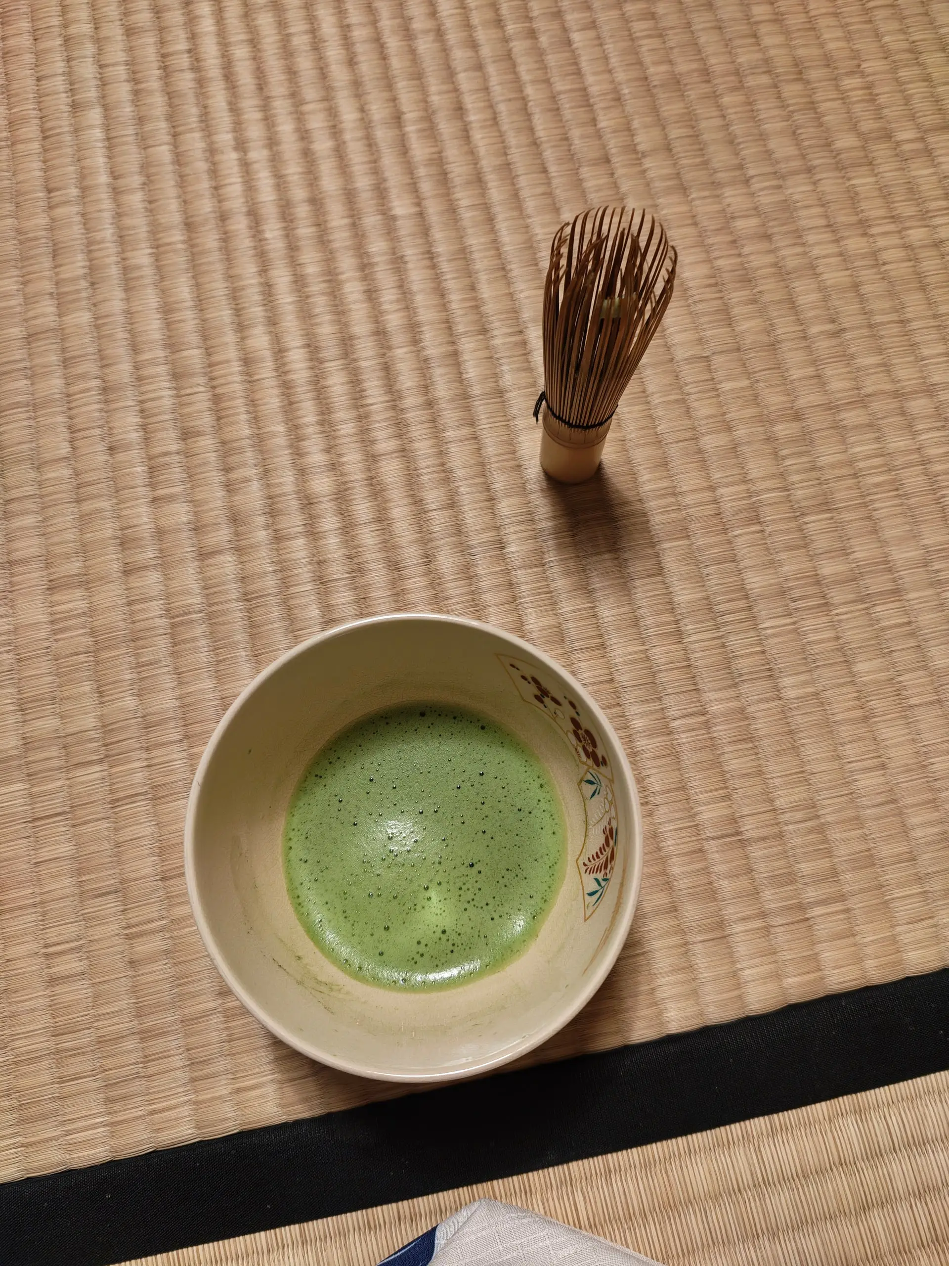 Miska napěněného zeleného matcha čaje a bambusový šlehač položené na tatami rohoži.