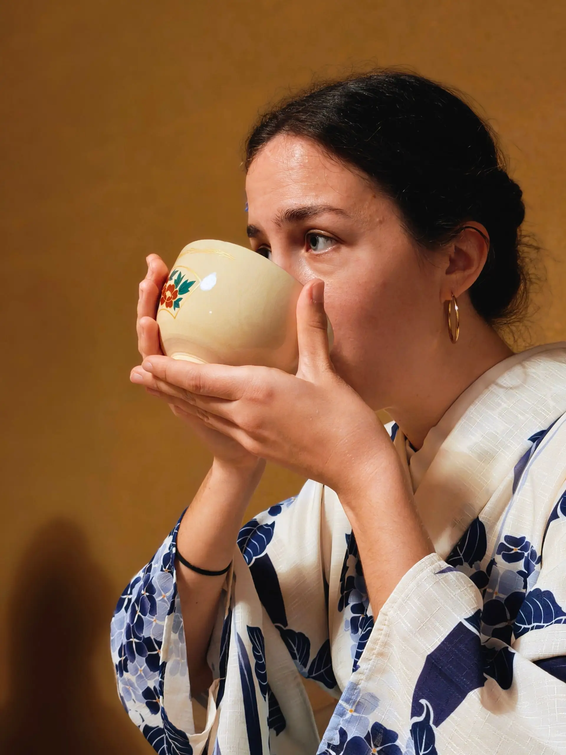 Žena v modrobílém kimonu drží oběma rukama keramickou misku a upíjí čaj.