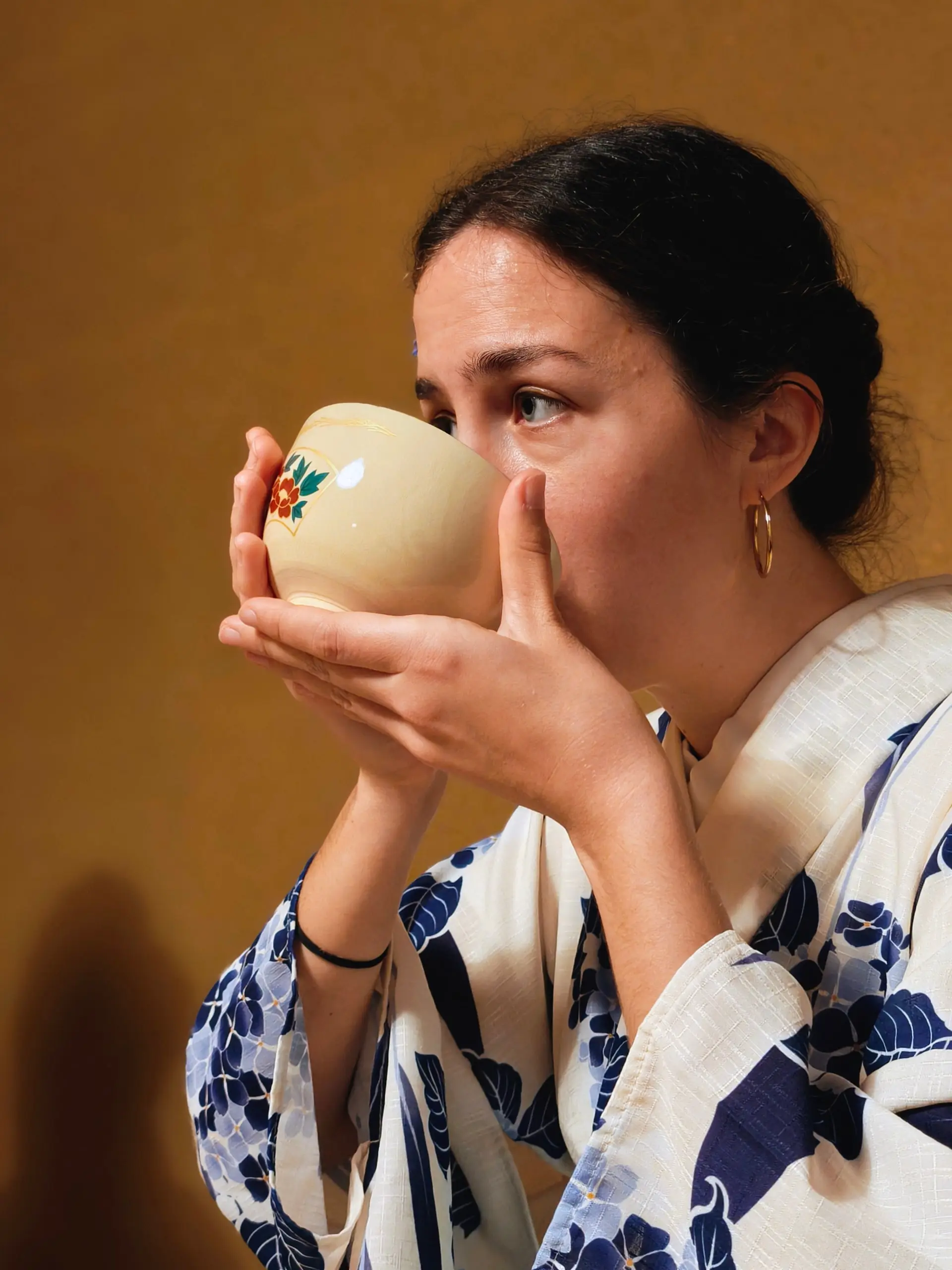 Žena v modrobílém kimonu drží oběma rukama keramickou misku a upíjí čaj.