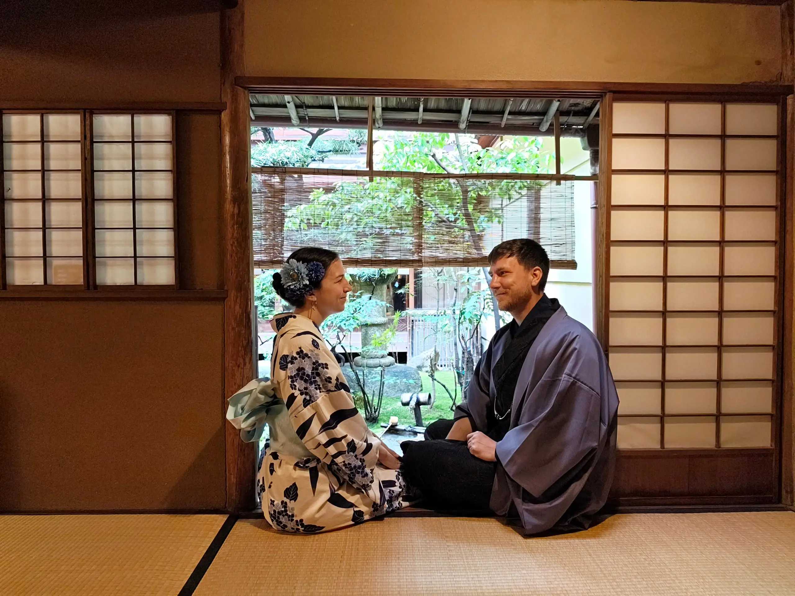 Žena a muž v kimonech sedí proti sobě na tatami u okna s výhledem na zenovou zahradu.