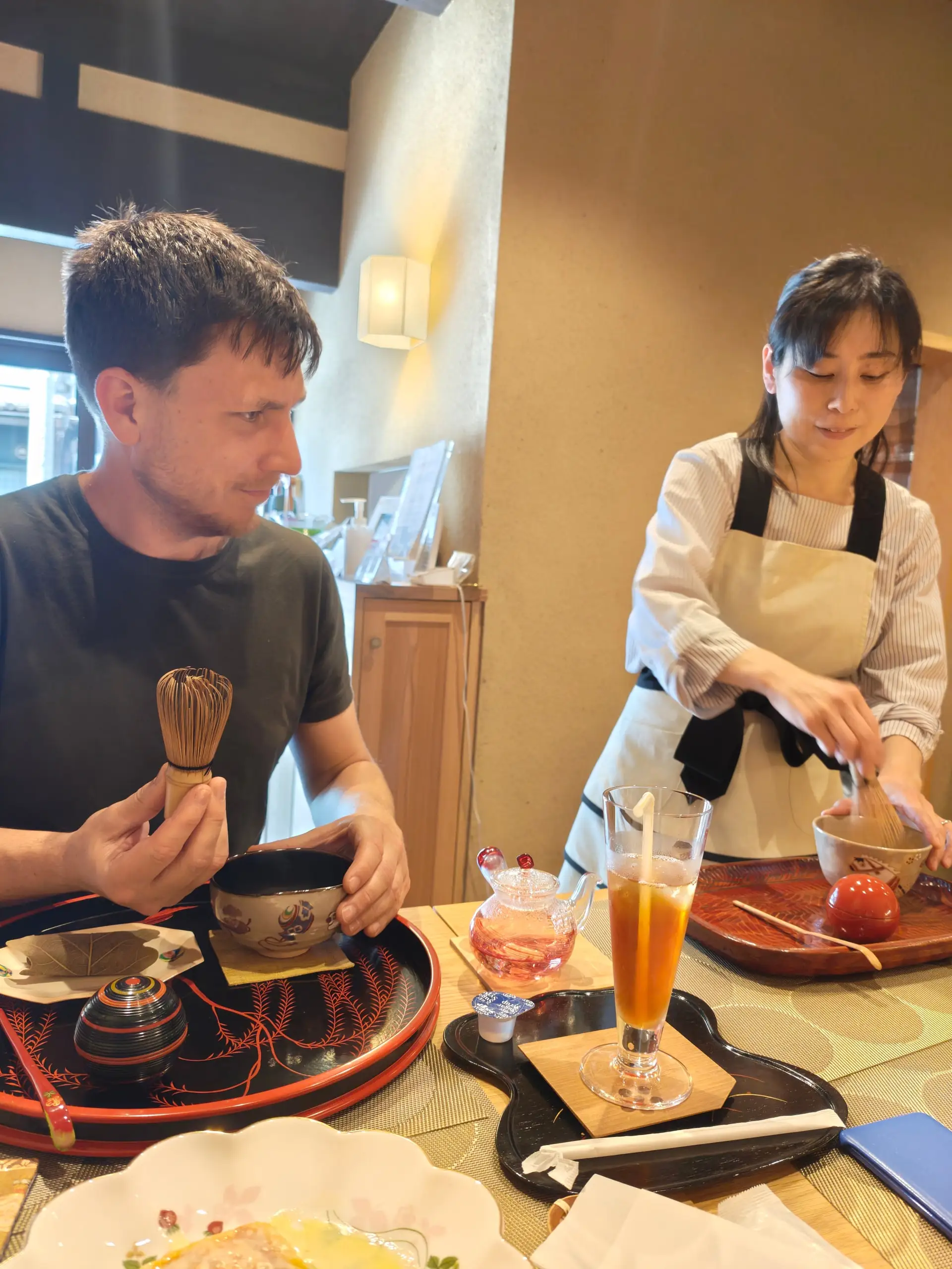 Muž šlehá matchu bambusovým štětcem zatímco japonská hostitelka v zástěře připravuje čaj u stolu.