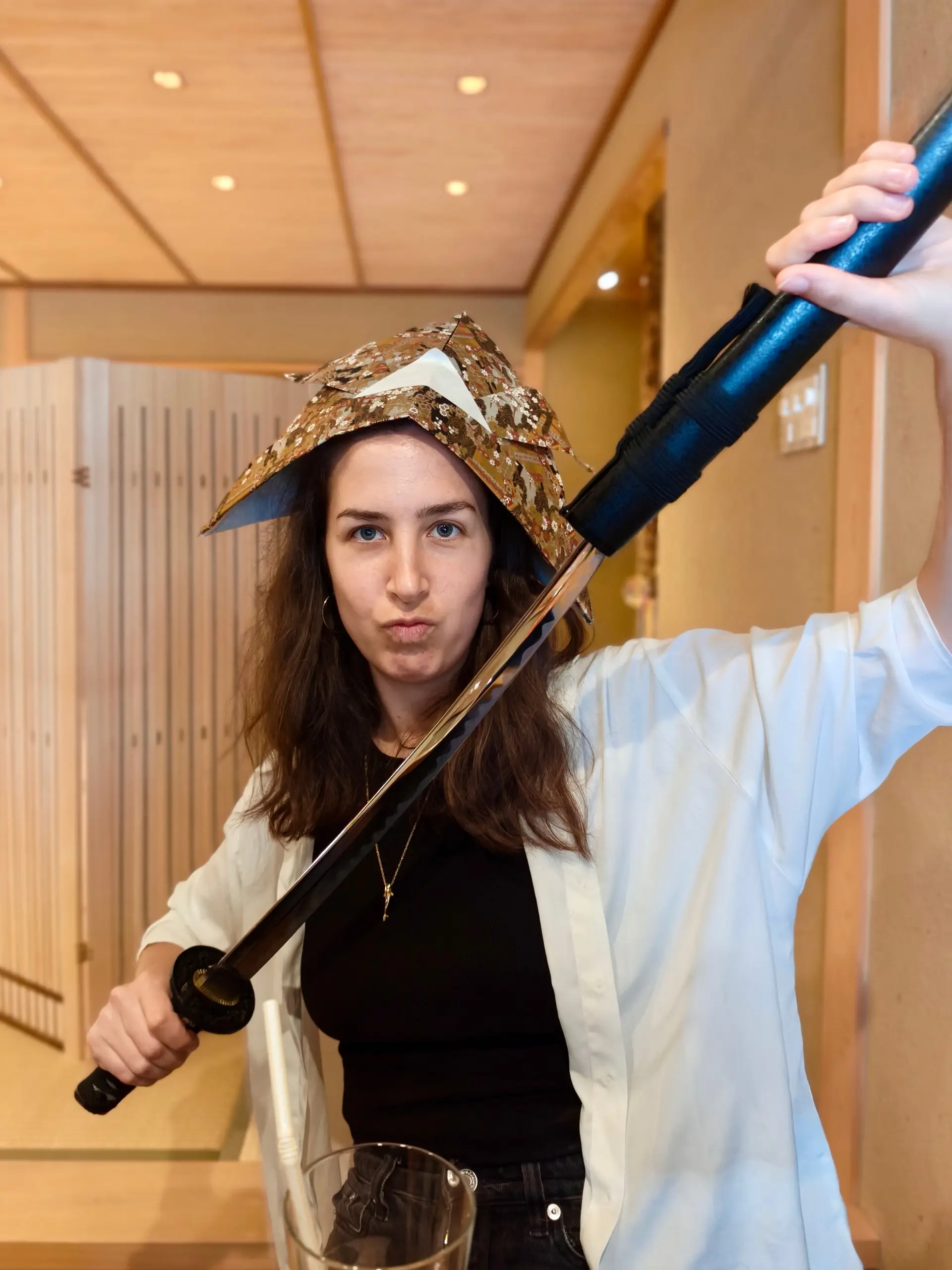 Žena s japonskou samurajskou čapkou na hlavě drží katanu, kterou jí někdo podává.