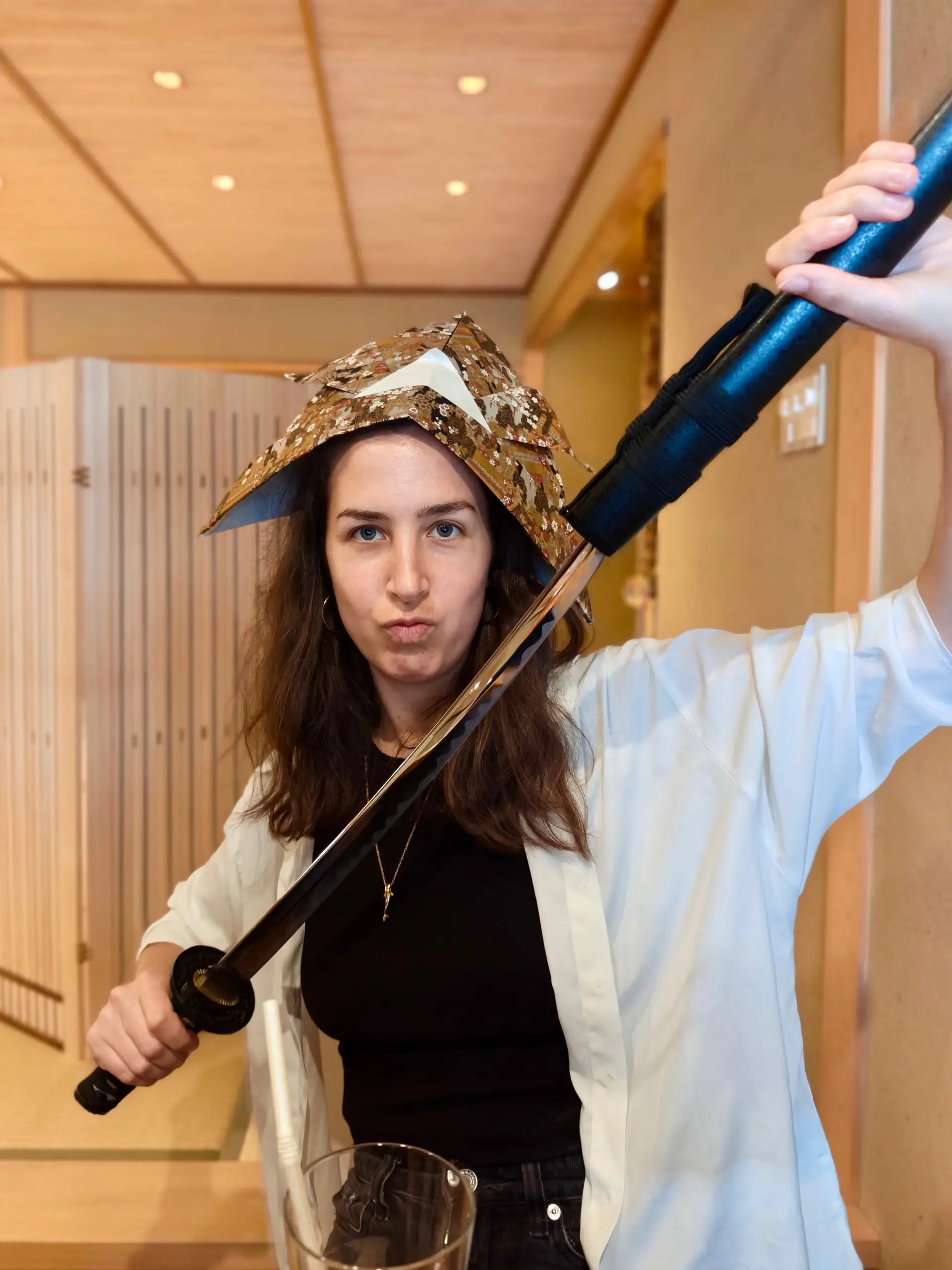 Žena s japonskou samurajskou čapkou na hlavě drží katanu, kterou jí někdo podává.