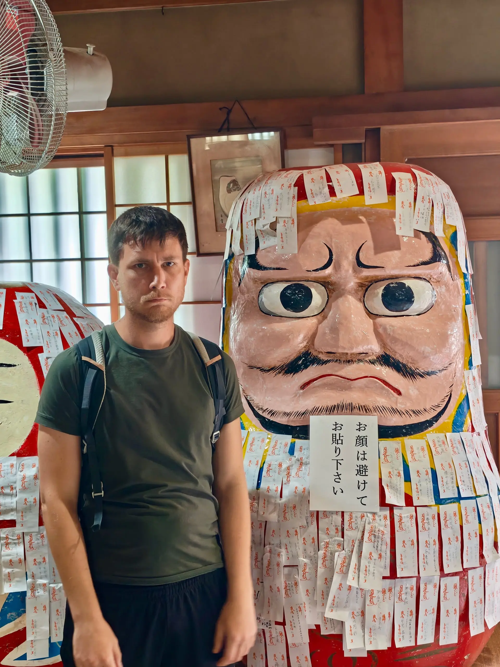 Muž s batohem stojí vedle obří malované sošky daruma polepené modlitebními lístky ve svatyni.