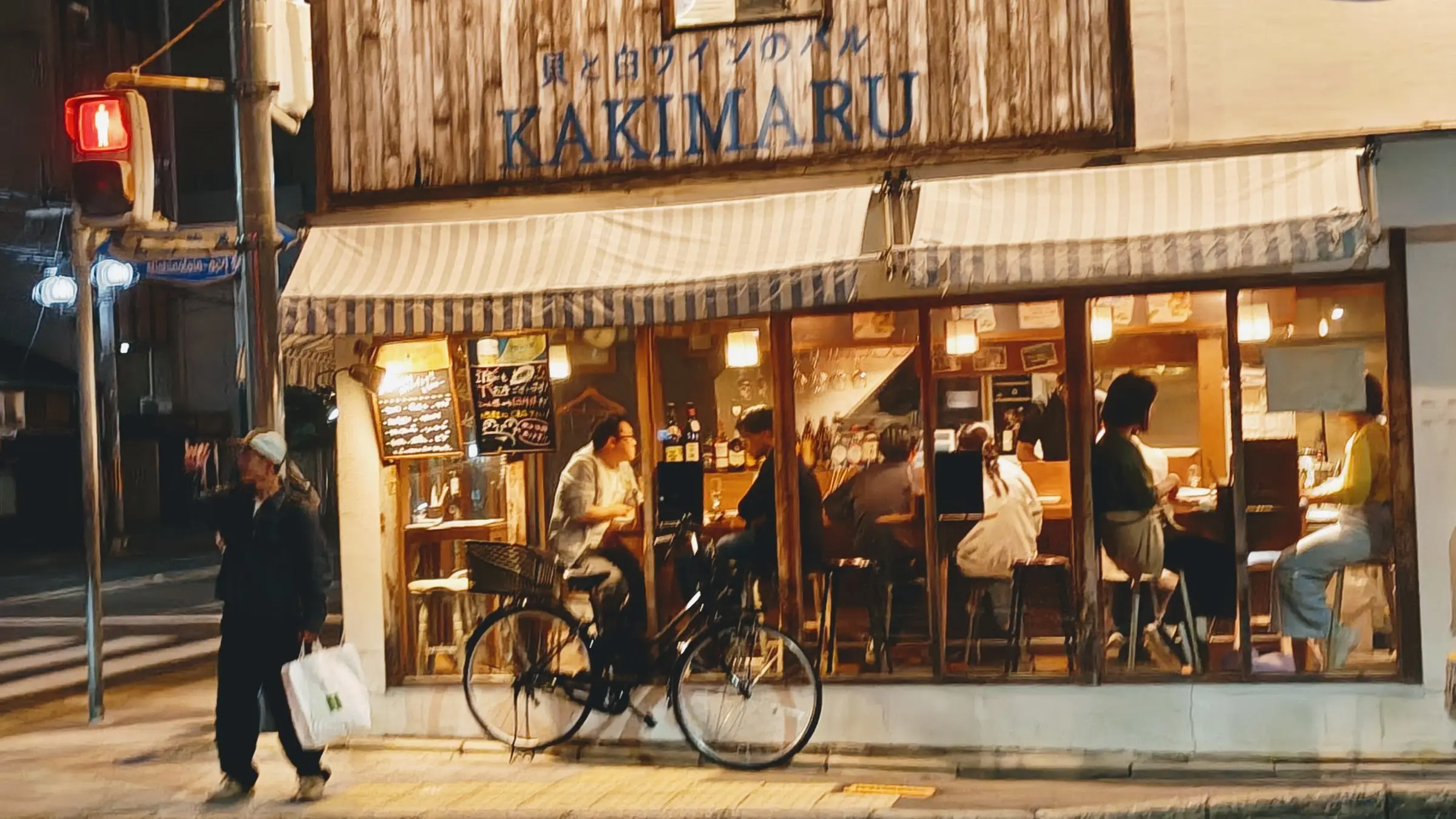 Osvětlený vinný bar Kakimaru s pruhovanou markýzou a hosty uvnitř za večerní ulicí s cyklistou.