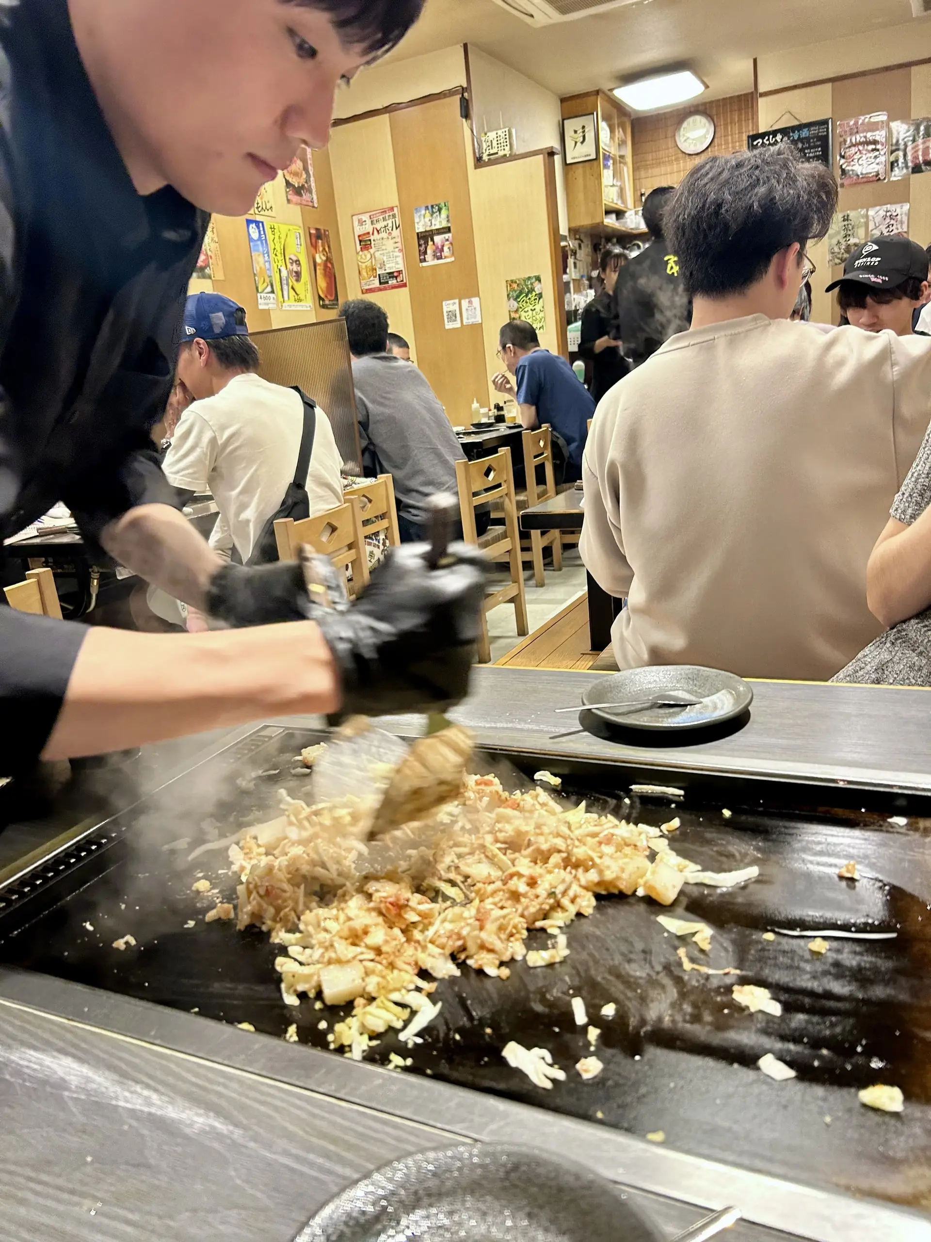 Kuchař v rukavici obrací a krájí japonské okonomiyaki na rozpálené plotně přímo u stolu v rušné tokijské restauraci.