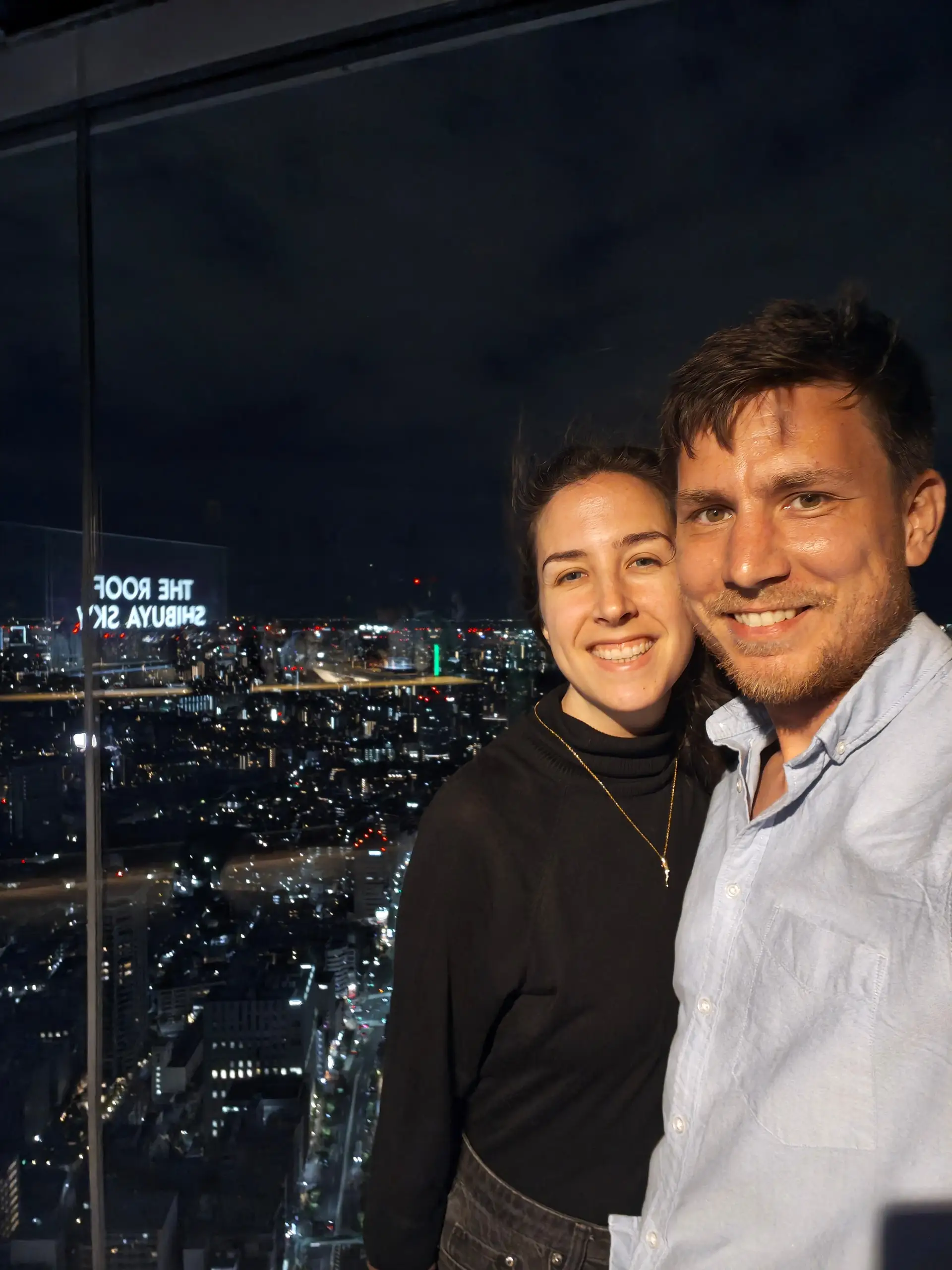 Pár se usmívá u prosklené stěny s panoramatem nočního Tokia za zády.