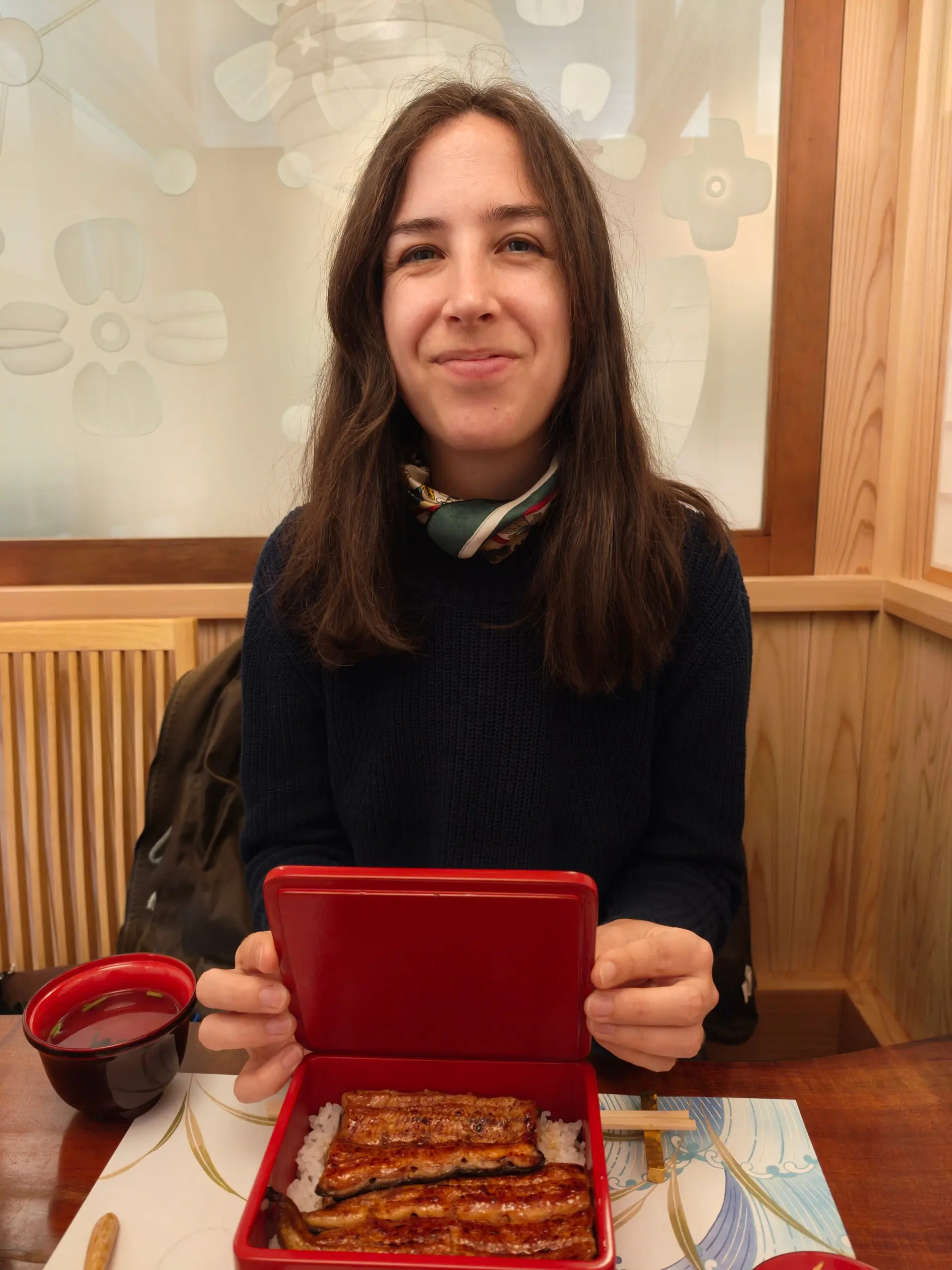 Žena u stolu drží červenou krabičku s grilovaným úhořem na rýži a polévkou.
