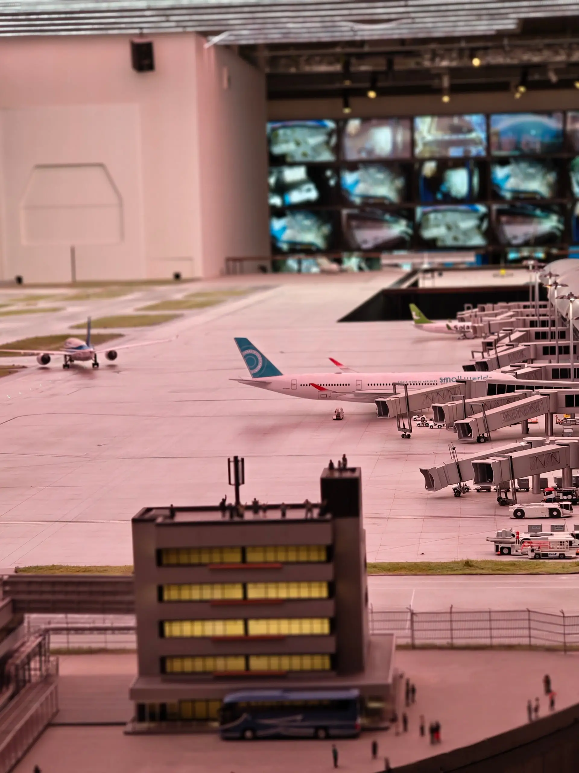 Miniaturní model letiště s letadly a stěnou monitorů na výstavě Small Worlds.