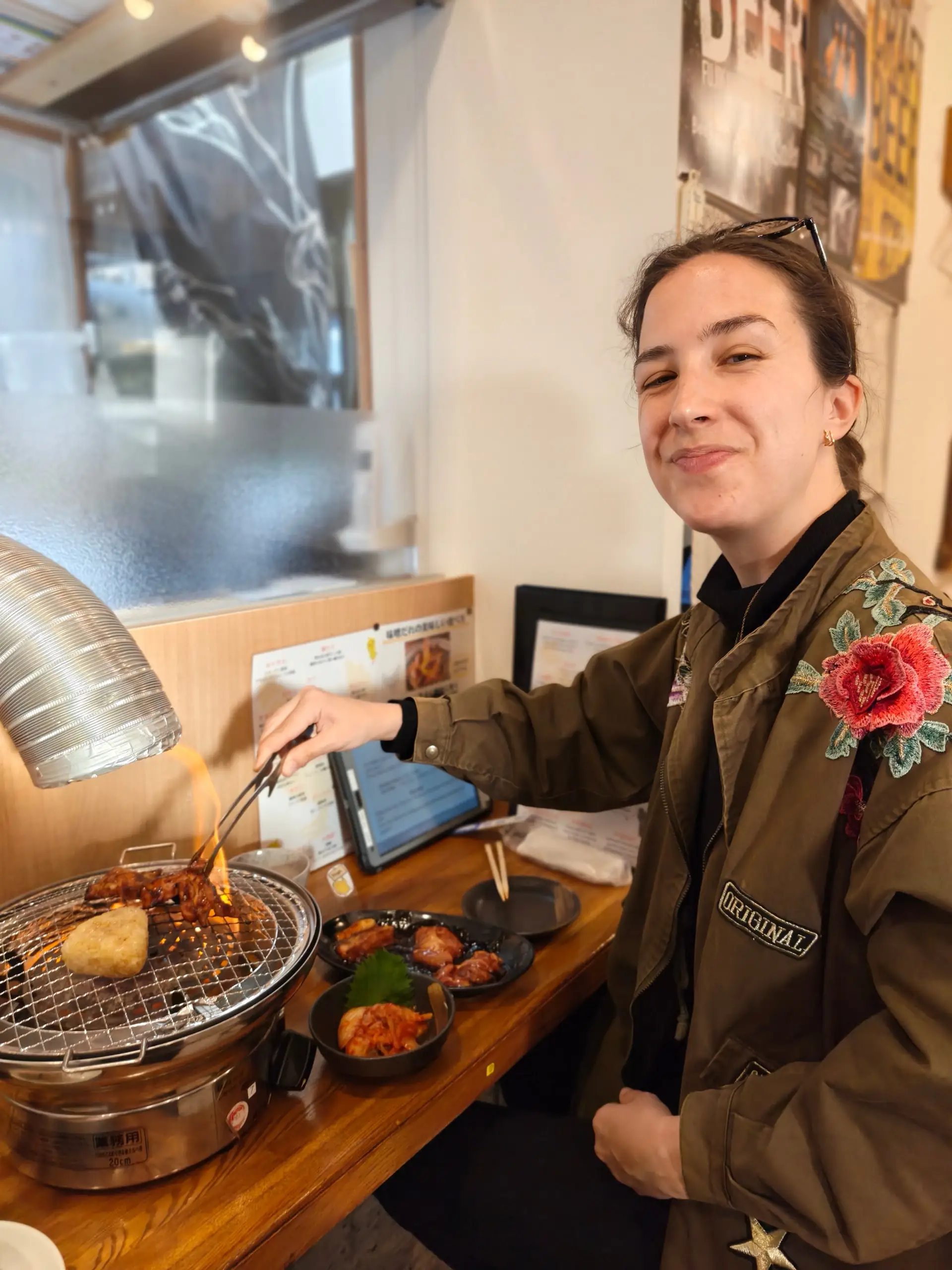 Usměvavá žena obrací jídlo na malém stolním grilu v restauraci s miskami pokrmů.