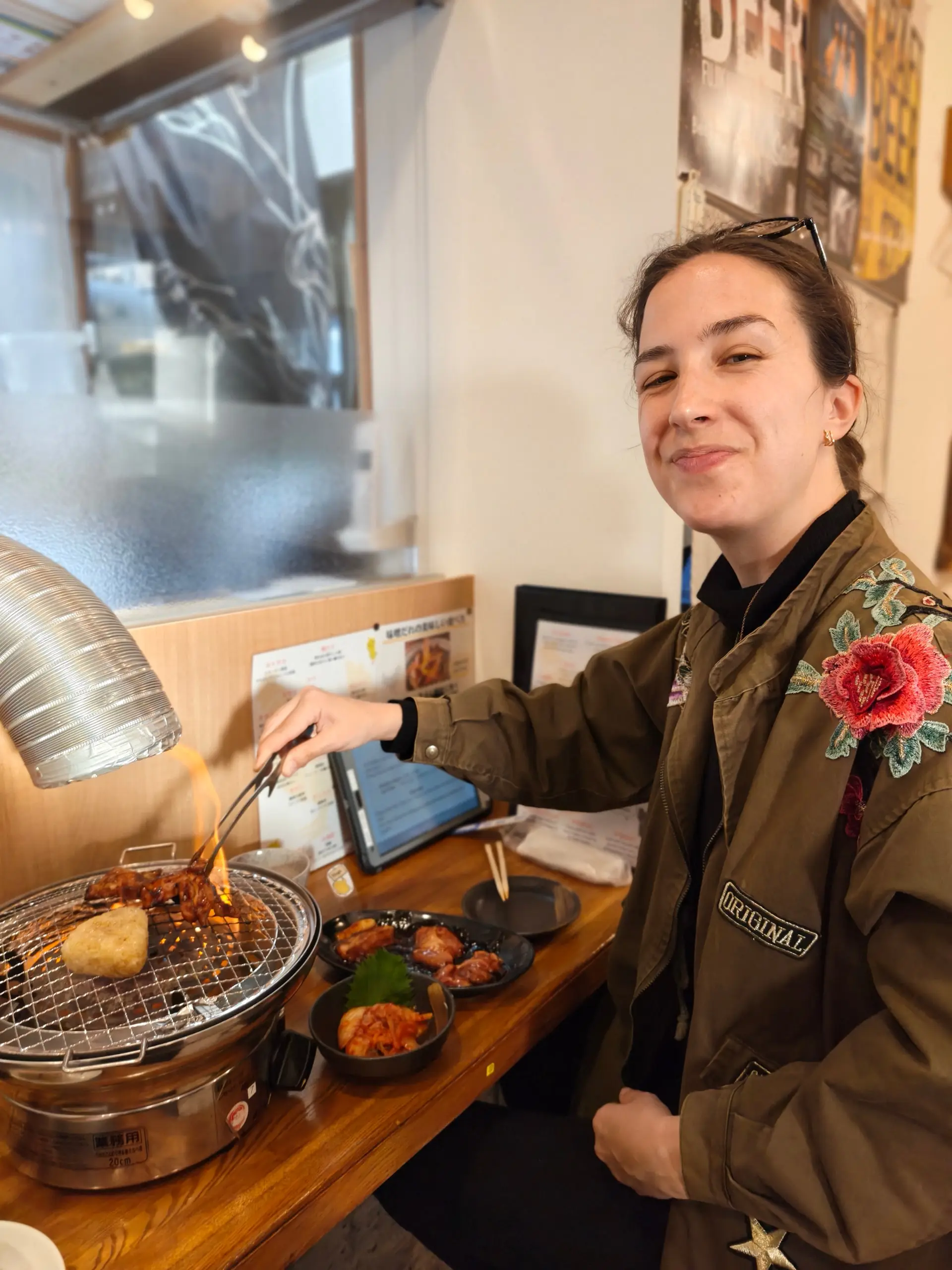 Usměvavá žena obrací jídlo na malém stolním grilu v restauraci s miskami pokrmů.