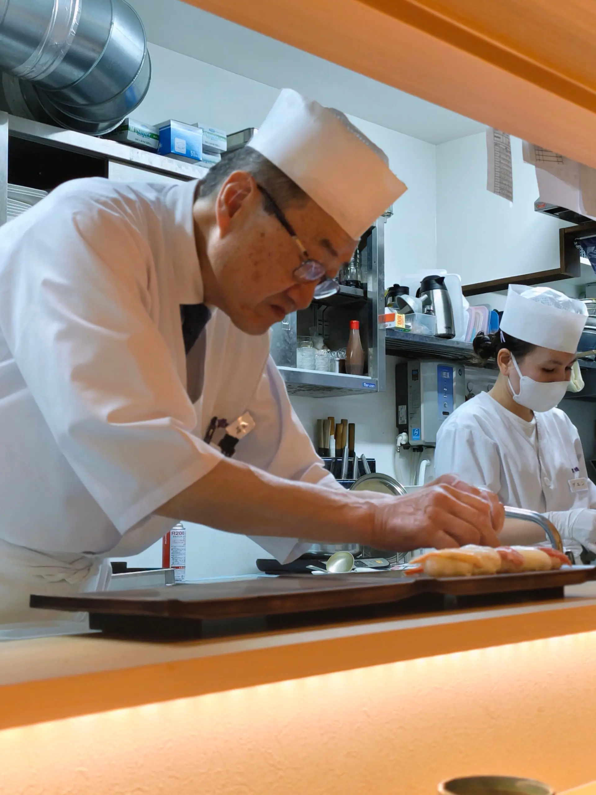 Sushi kuchař v bílém připravuje nigiri za pultem restaurace, vzadu druhá kuchařka.