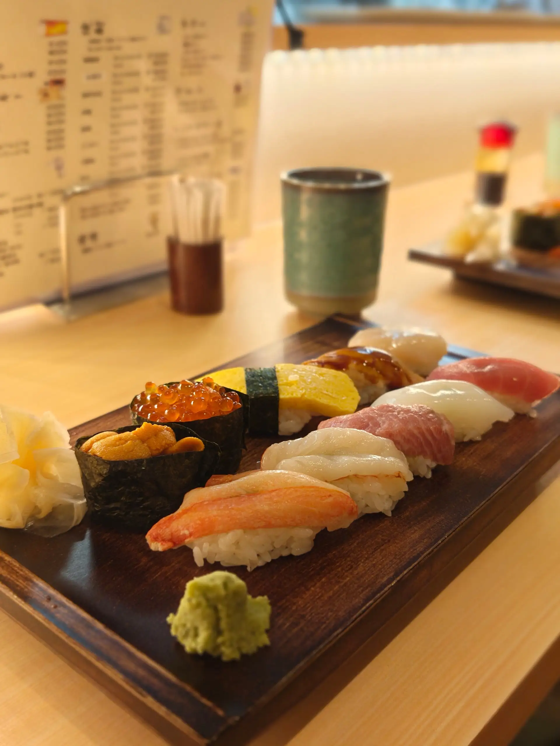 Dřevěný talíř s deseti druhy nigiri sushi, wasabi a nakládaným zázvorem na stole.