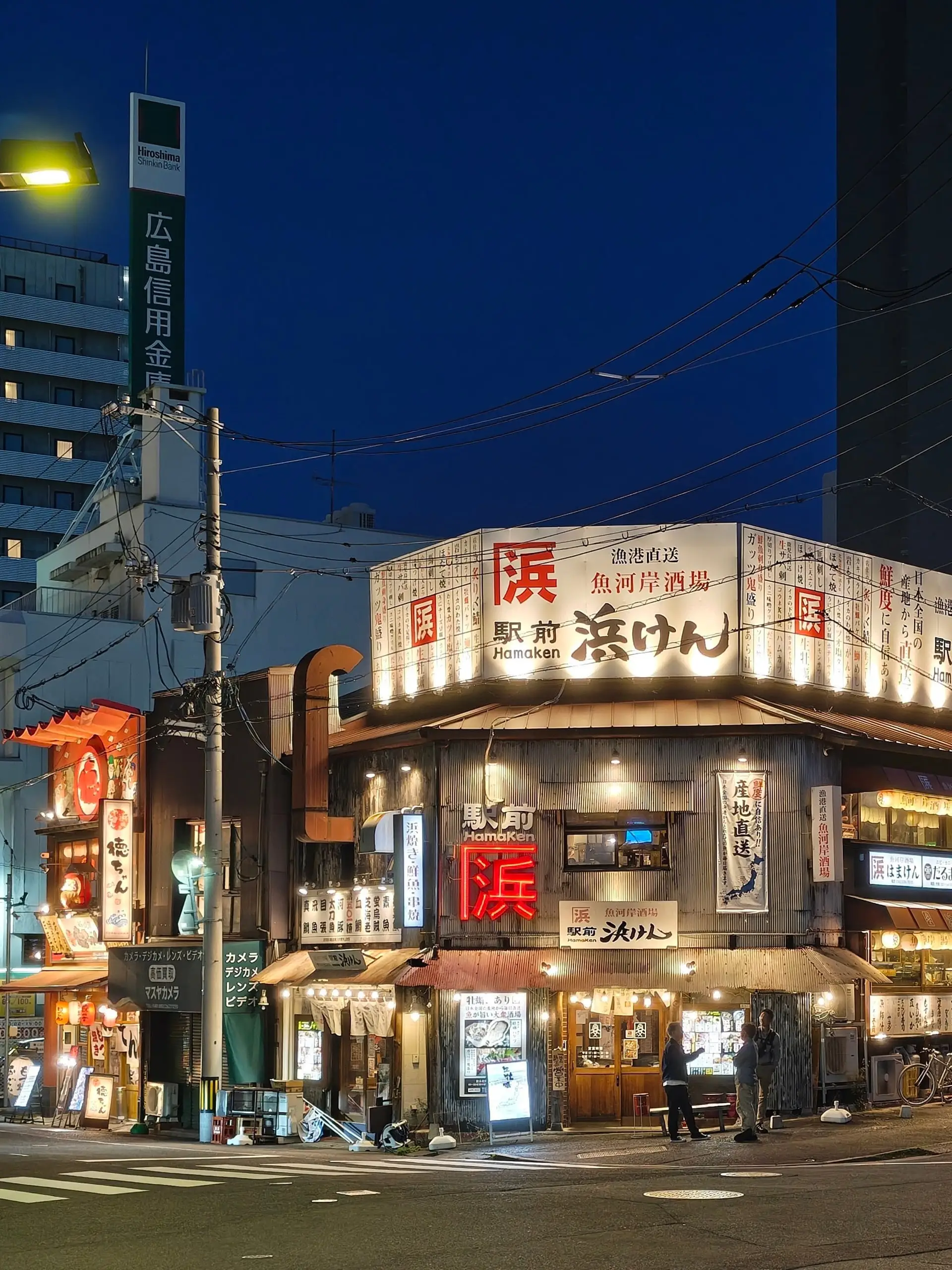 Rohová japonská restaurace s rozsvícenými červenými nápisy a lampiony za modré večerní oblohy.