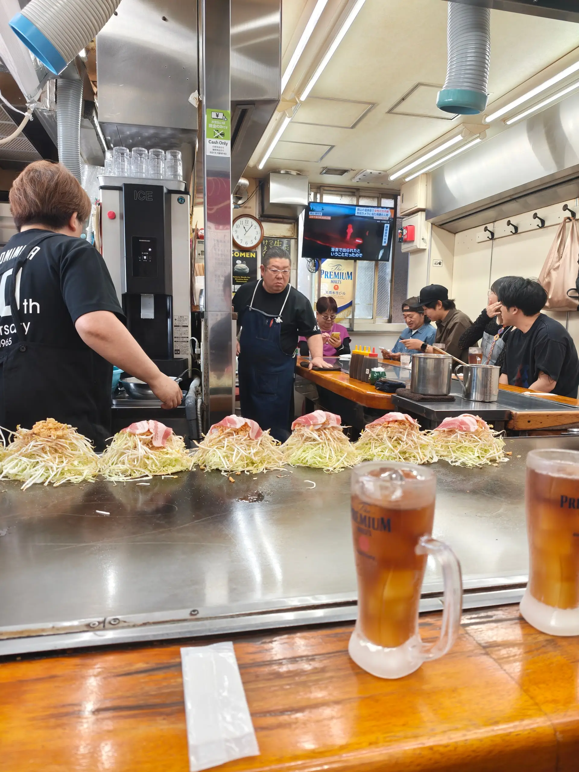 Kuchaři smaží na velké plotně řady okonomiyaki se zelím a slaninou, vpředu dva korbele.