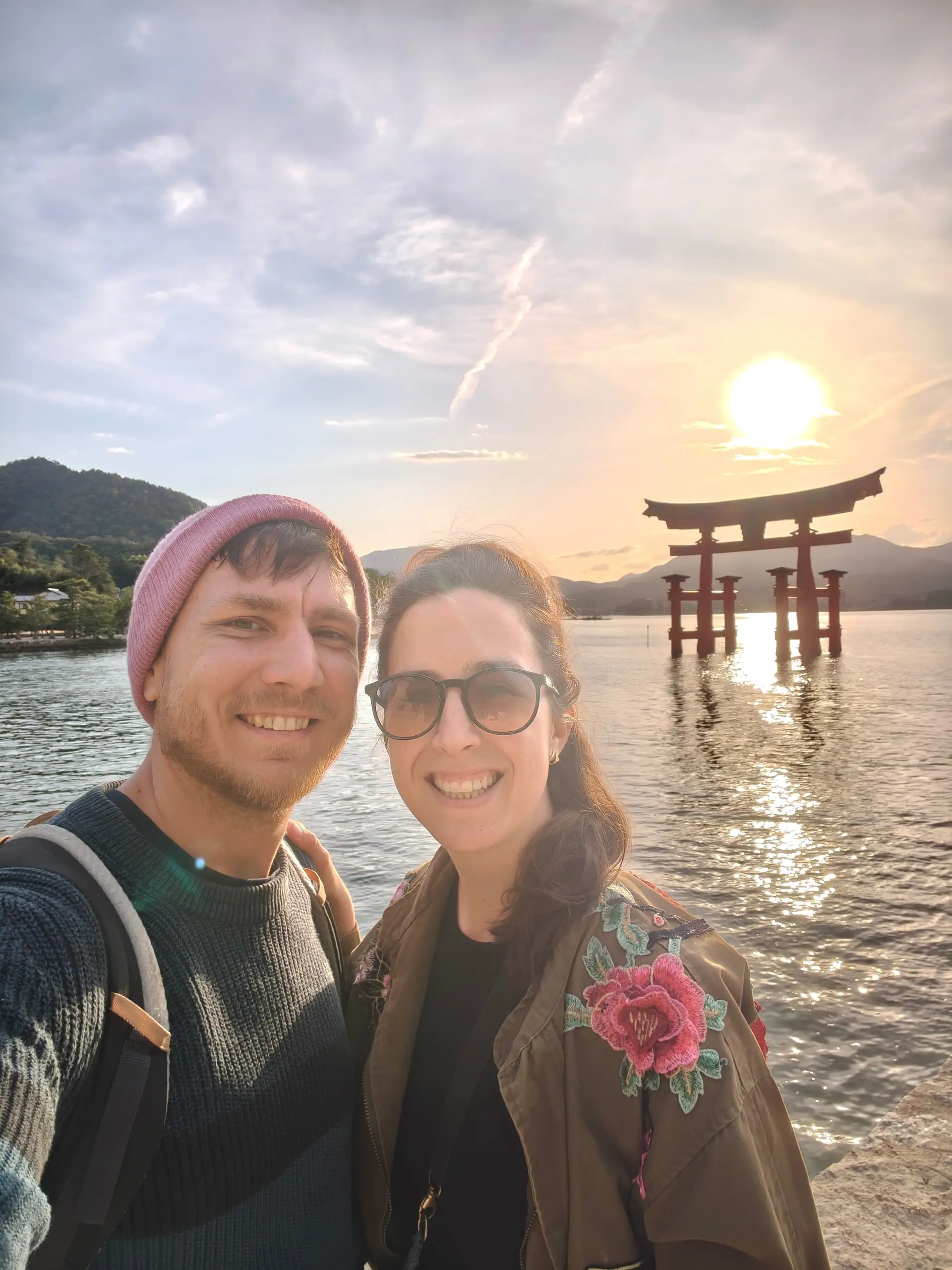 Usměvavý pár v selfie u moře, v pozadí brána torii a zapadající slunce.