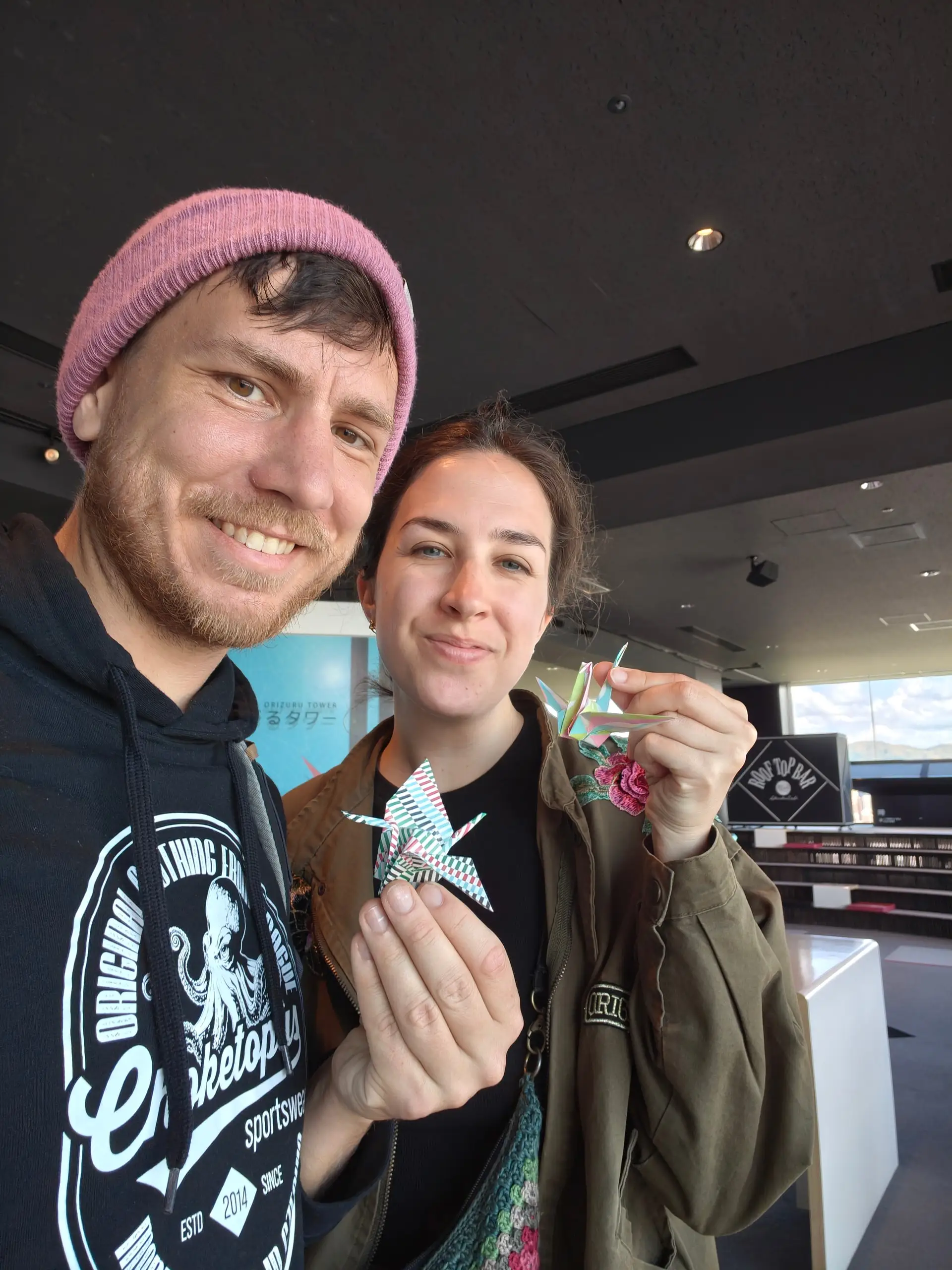 Usměvavý pár drží barevné papírové jeřáby origami na prosklené vyhlídce.